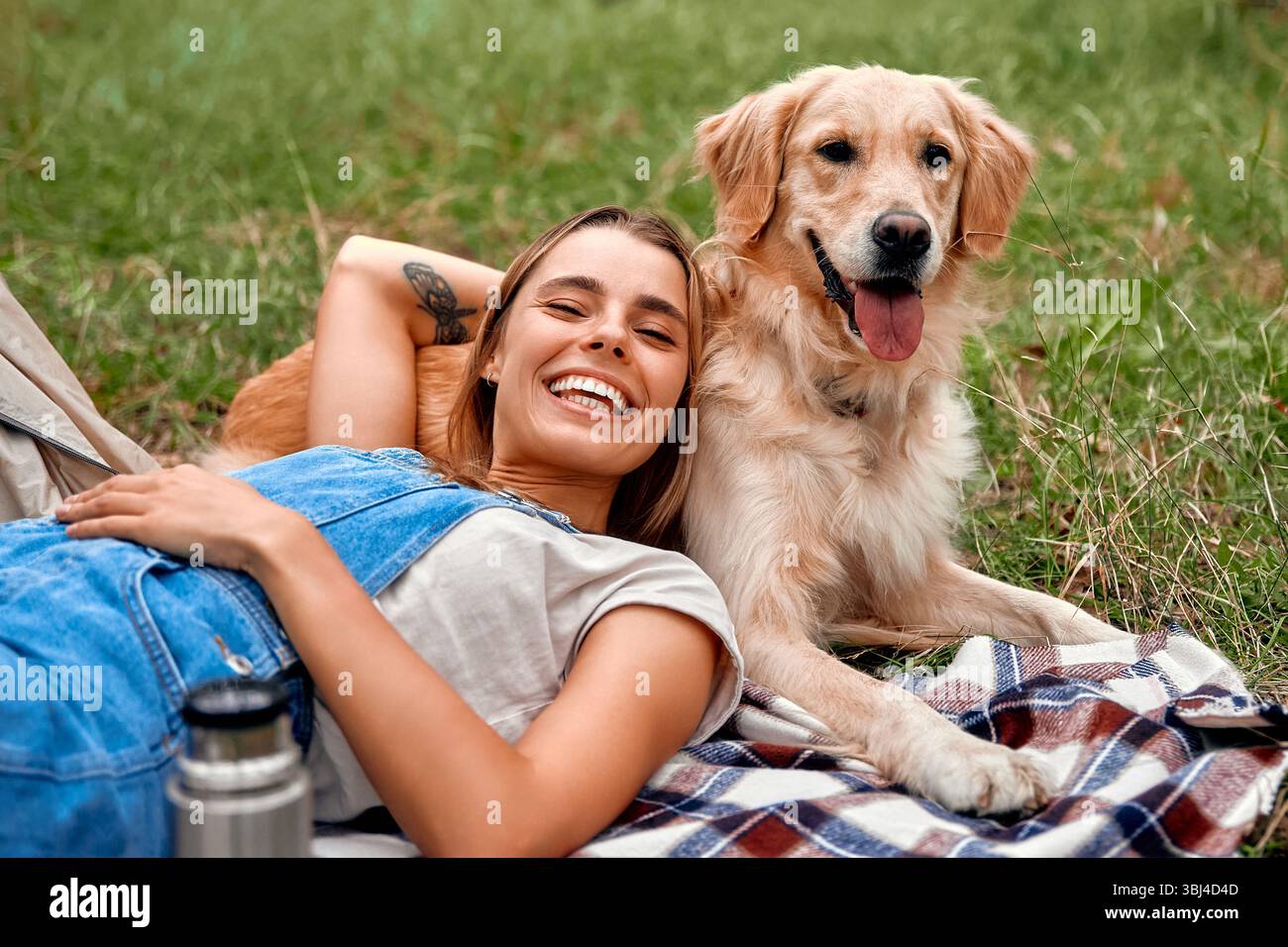 Eine fröhliche Frau ist völlig in den Genuss eines schönen sonnigen Tages eingetaucht, während sie mit ihrem verspielten goldenen Retriever, captain, bequem auf dem üppigen Gras liegt Stockfoto