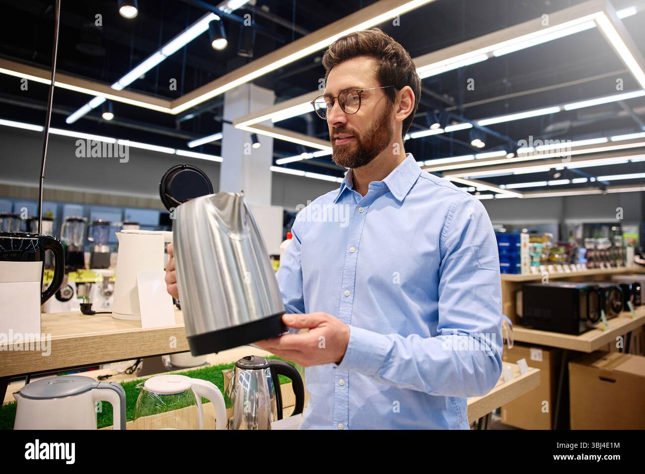 Ein Mann untersucht sorgfältig einen eleganten, glänzenden Wasserkocher aus Edelstahl, während er von einer Vielzahl moderner Küchengeräte in einem stilvollen Einzelhandelsbereich des umgeben ist Stockfoto