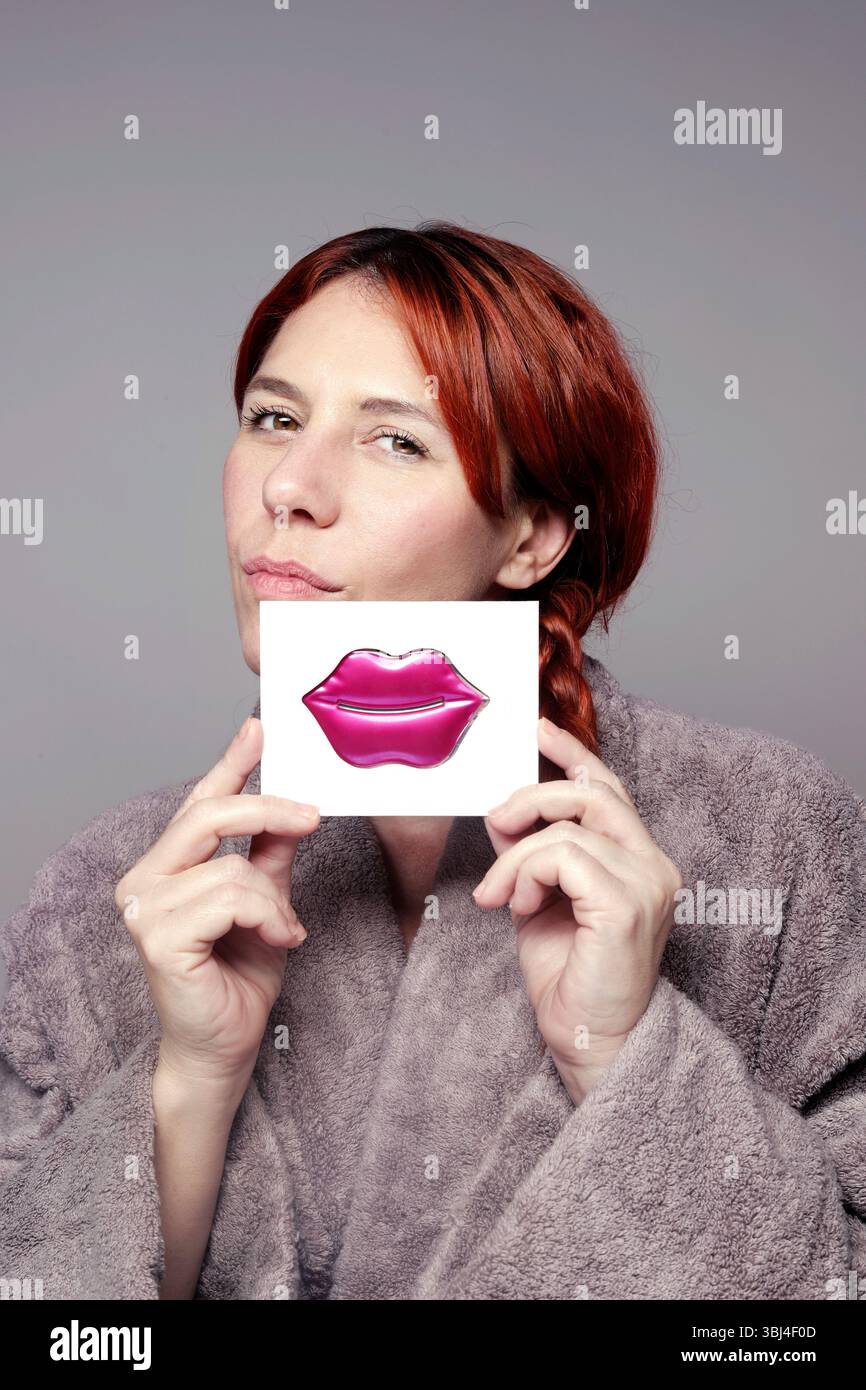 Rothaarige Frau, die einen Bademantel trägt und einen lippenförmigen Schönheitsbehandlungsstreifen vor ihrem Gesicht hält Stockfoto