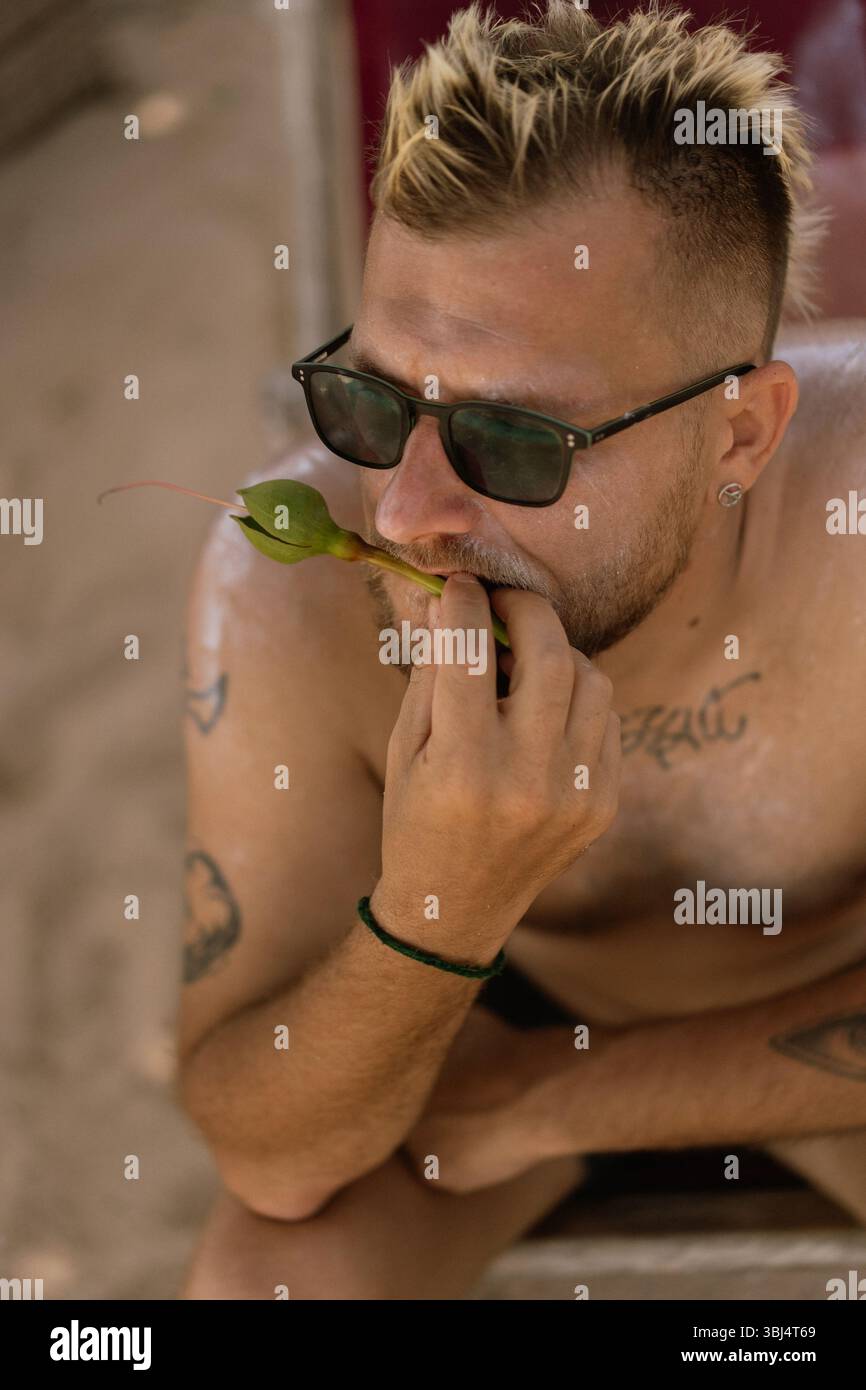 Tätowierter Mann in Sonnenbrille, der auf tropischen Pflanzenstamm am Strand beißt Stockfoto