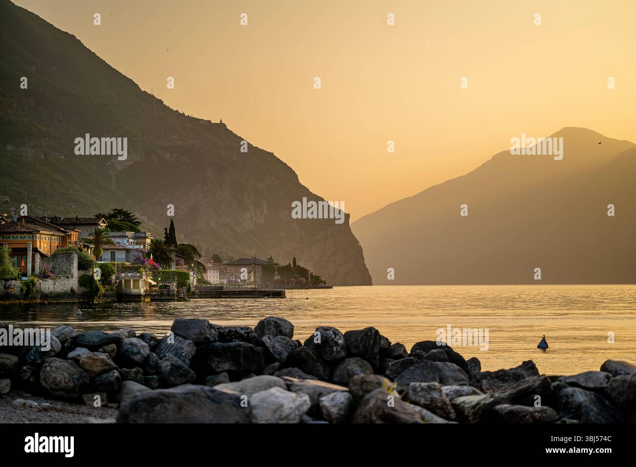 Farbenfroher Sonnenaufgang über dem Gardasee in Italien, mit ruhigem Seewasser im Vordergrund und trüben Bergen im Hintergrund, die eine friedliche, ruhige Atmosphäre schaffen. Stockfoto
