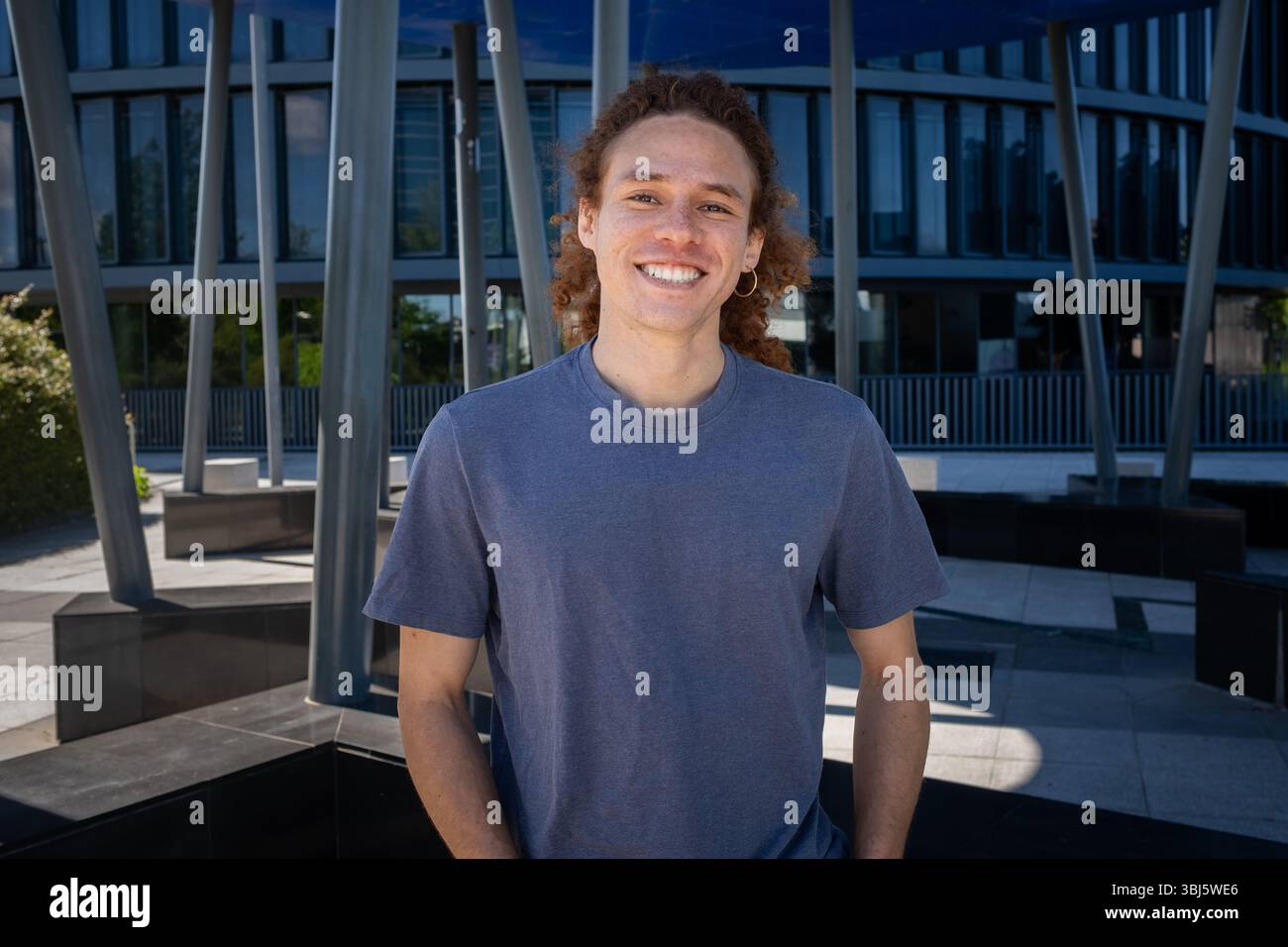 Junger Mann mit lockigen Haaren und Ohrringen, der draußen in moderner städtischer Umgebung lächelt. Lässige Mode, moderner Lifestyle, selbstbewusstes männliches Porträt. Stockfoto