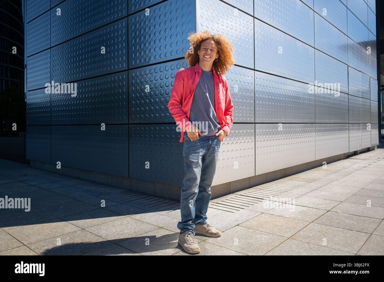 Junger Mann mit lockigen Haaren und rosa Jacke, der draußen vor einem modernen Metallgebäude steht, urbanes Mode- und Studentenleben-Porträt, Zukunft Stockfoto