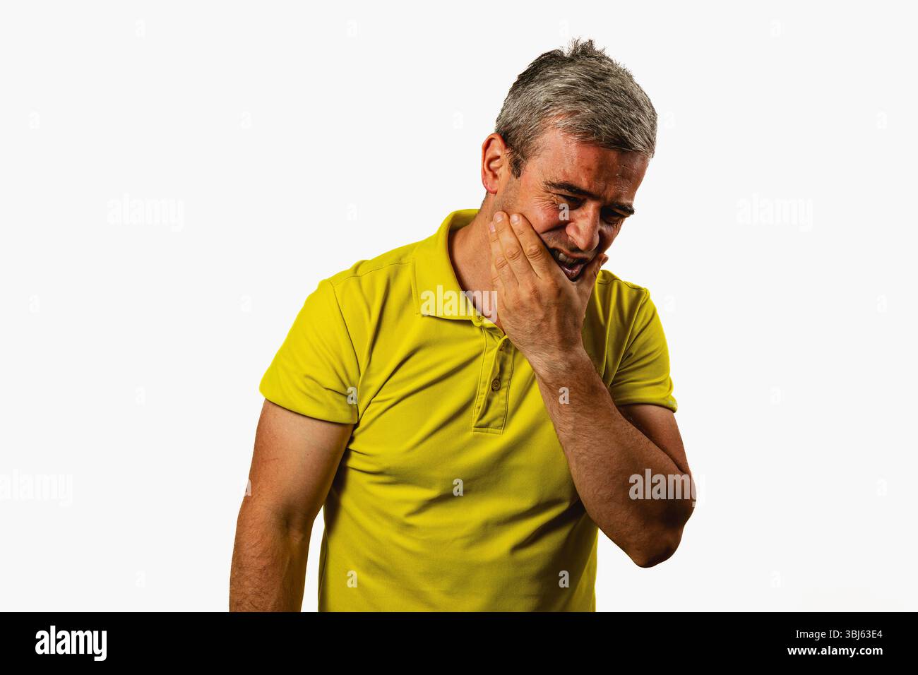 Ein Mann in einem gelben Hemd zeigt Anzeichen von Unbehagen, wenn er drinnen vor einem schlichten Hintergrund steht, was einen Moment des Unbehagens oder der Not reflektiert. Stockfoto