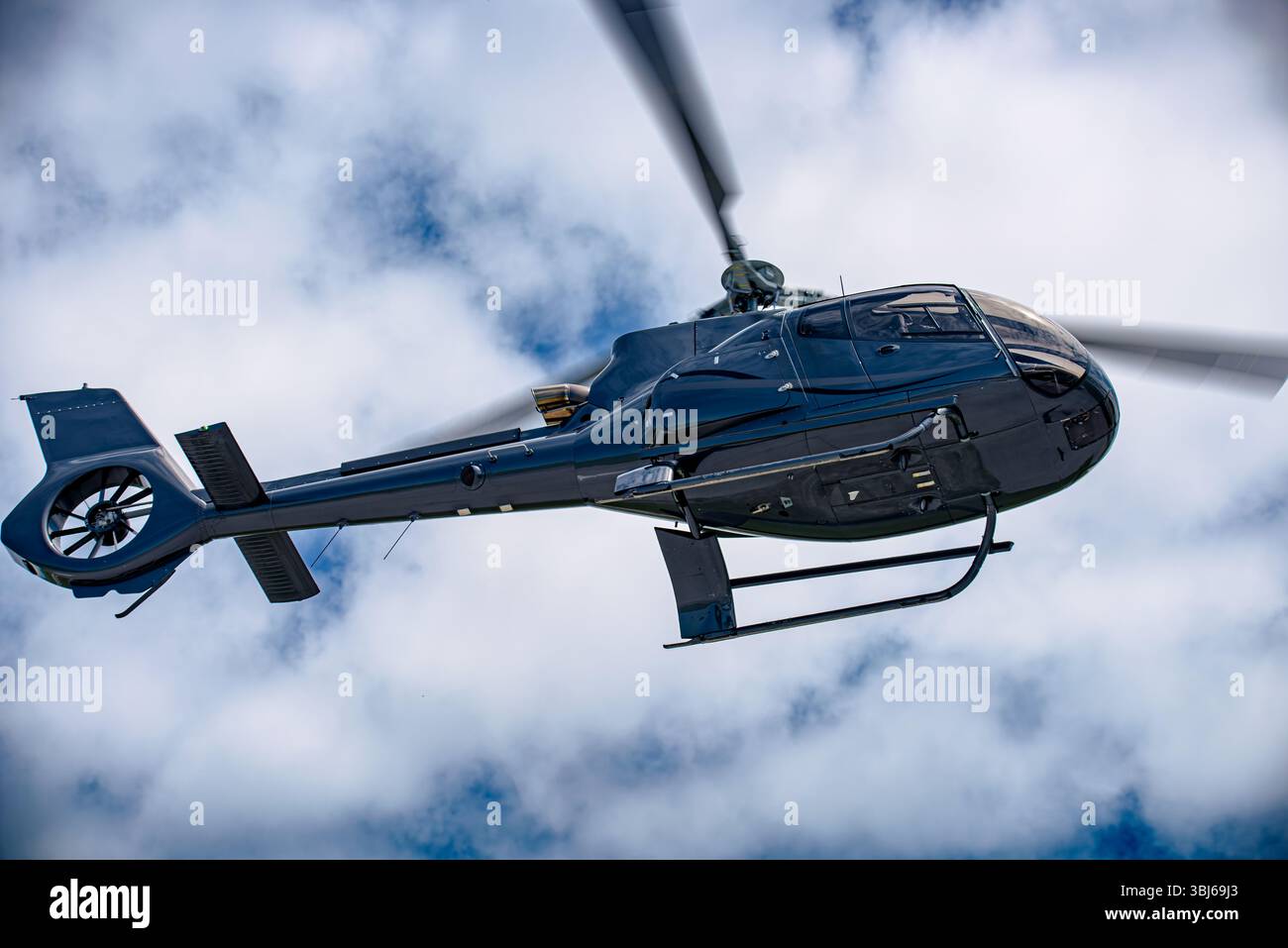 Moderner schwarzer Hubschrauber im Flug gegen teilweise bewölkten Himmel, von unten fotografiert. Schlankes Design und geschlossener Heckrotor sichtbar. Stockfoto
