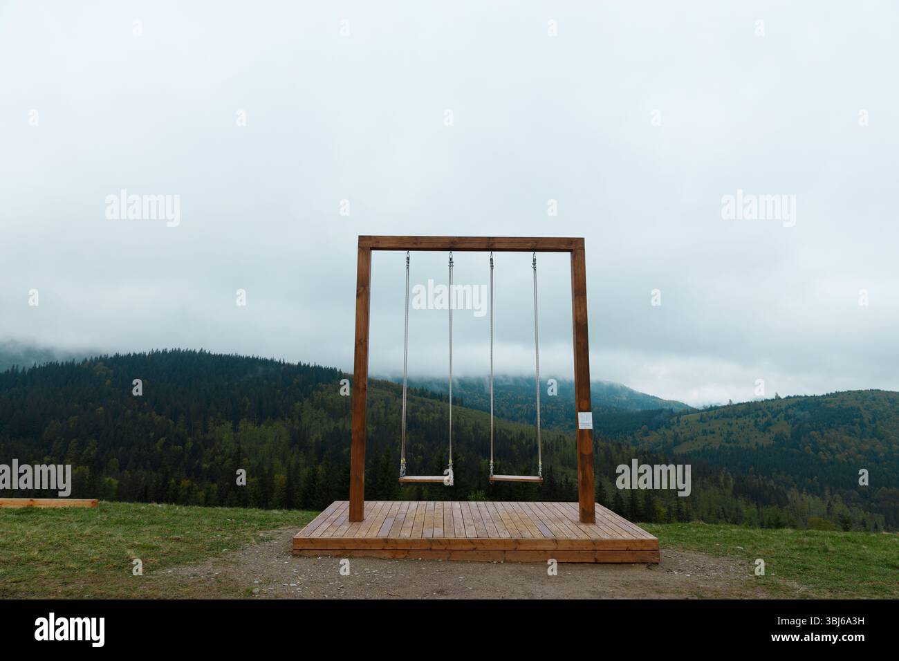Dieses Bild zeigt eine wunderschön gefertigte Holzschaukel vor der Kulisse von sanften grünen Hügeln und einem bewölkten Himmel, die Besucher zum Genießen von Tram einlädt Stockfoto