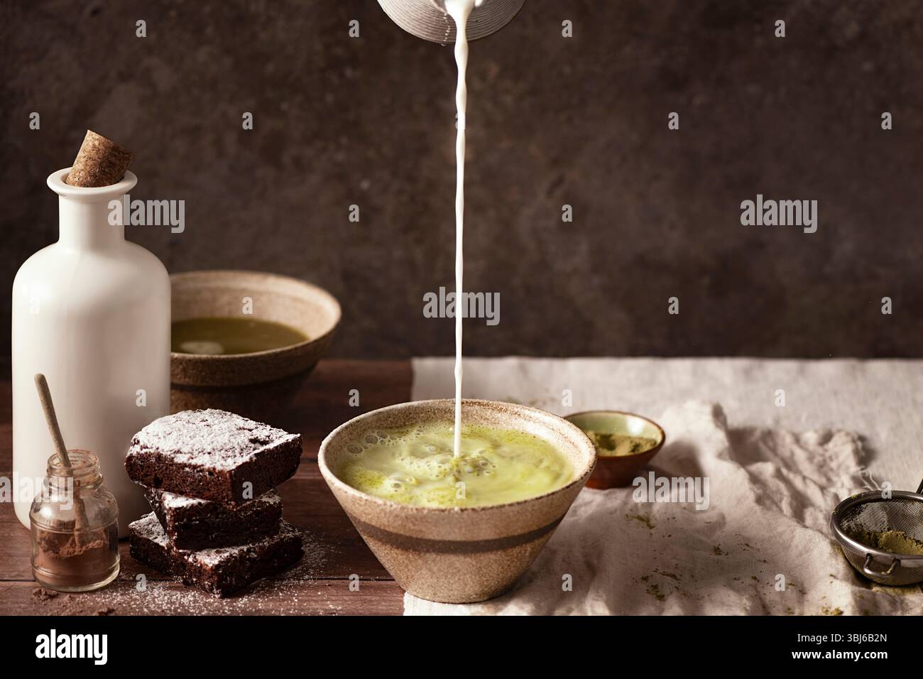 Zwei Schüsseln Matcha-Tee mit Brownies mit Kakaopulver auf dem Tisch, Milch wird in die Schüssel gegossen Stockfoto