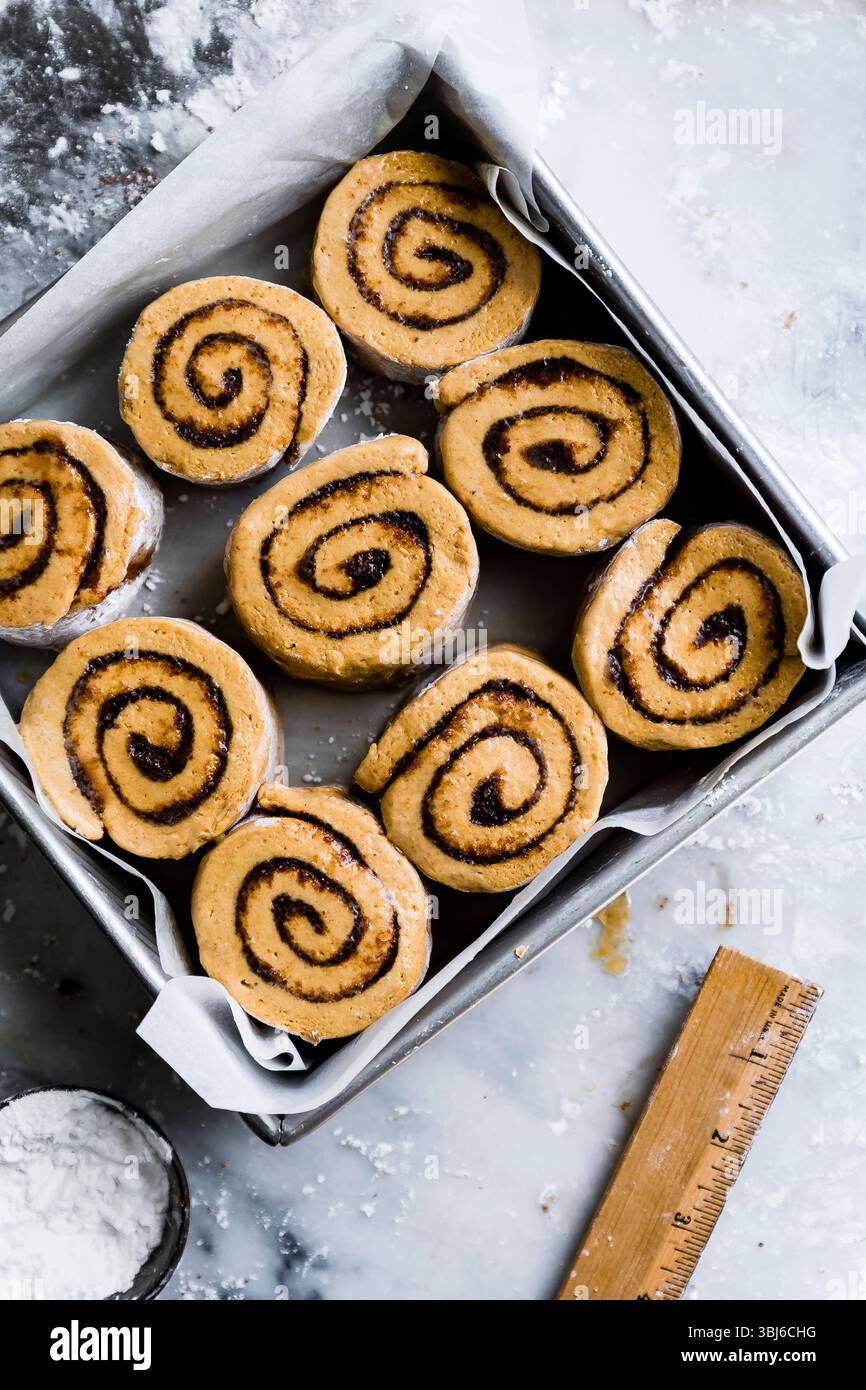 Ein Blick von oben auf ungebackene Zimtrollen, die in einem Backblech mit Backpapier auf einer bemehlten Oberfläche angeordnet sind. Stockfoto