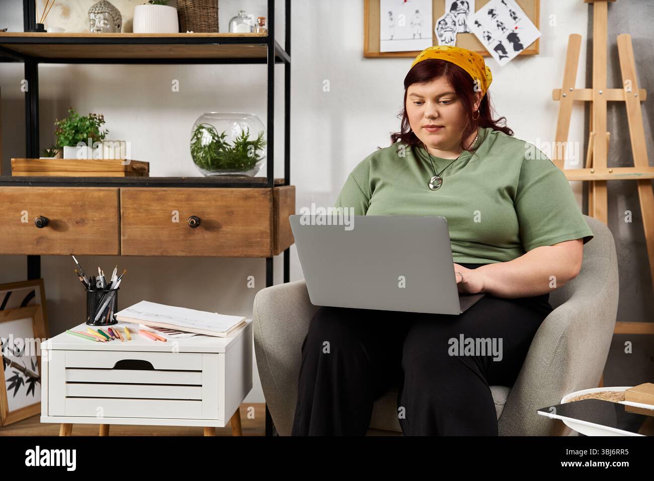 Frau arbeitet nachdenklich an einem Laptop in einem schicken Studio, das mit Kunstbedarf und Skizzen gefüllt ist Stockfoto