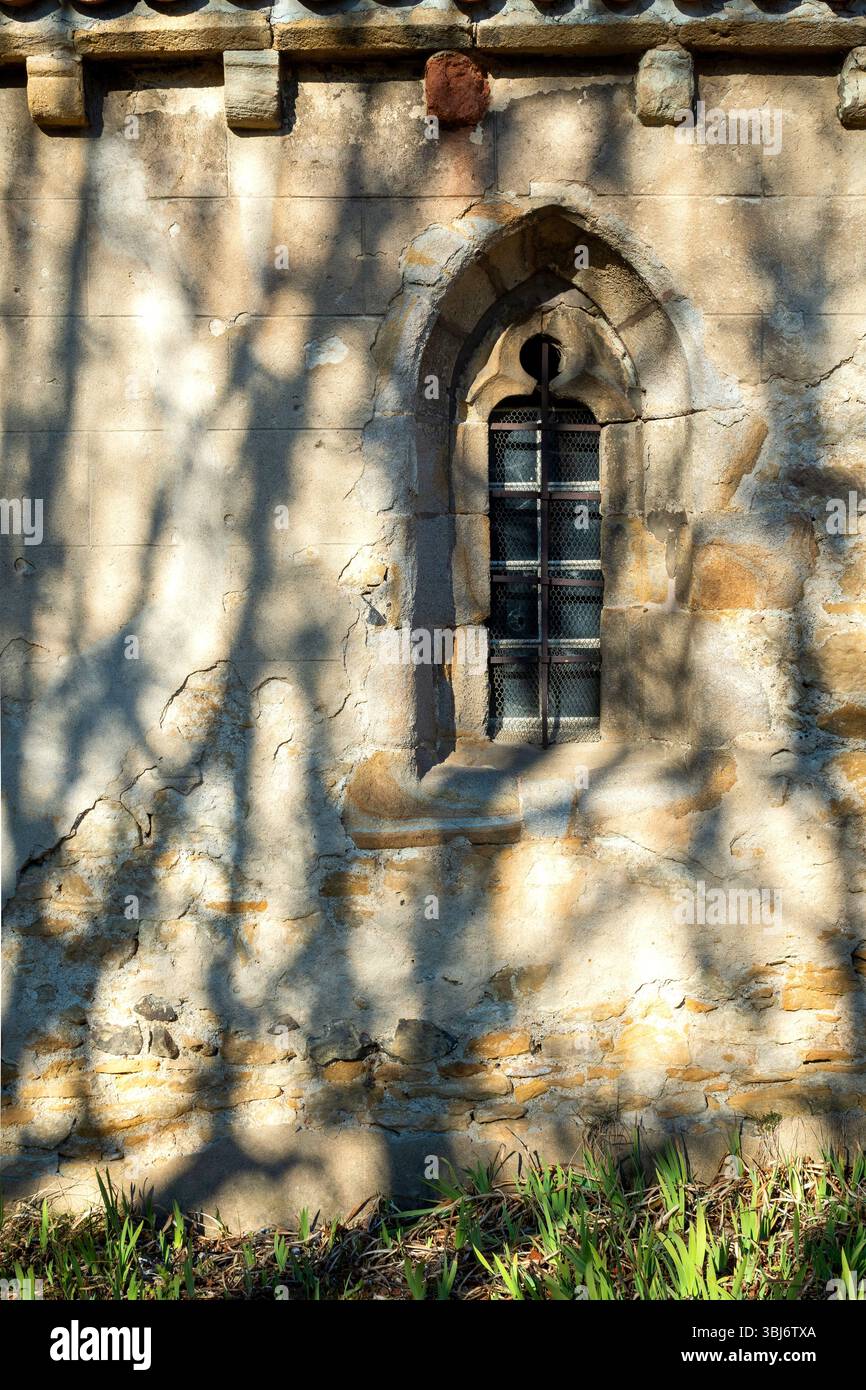 Schatten von umliegenden Bäumen bilden ein markantes Muster vor einer verwitterten Steinmauer mit einem kleinen Bogenfenster einer Kirche. . Frankreich Stockfoto