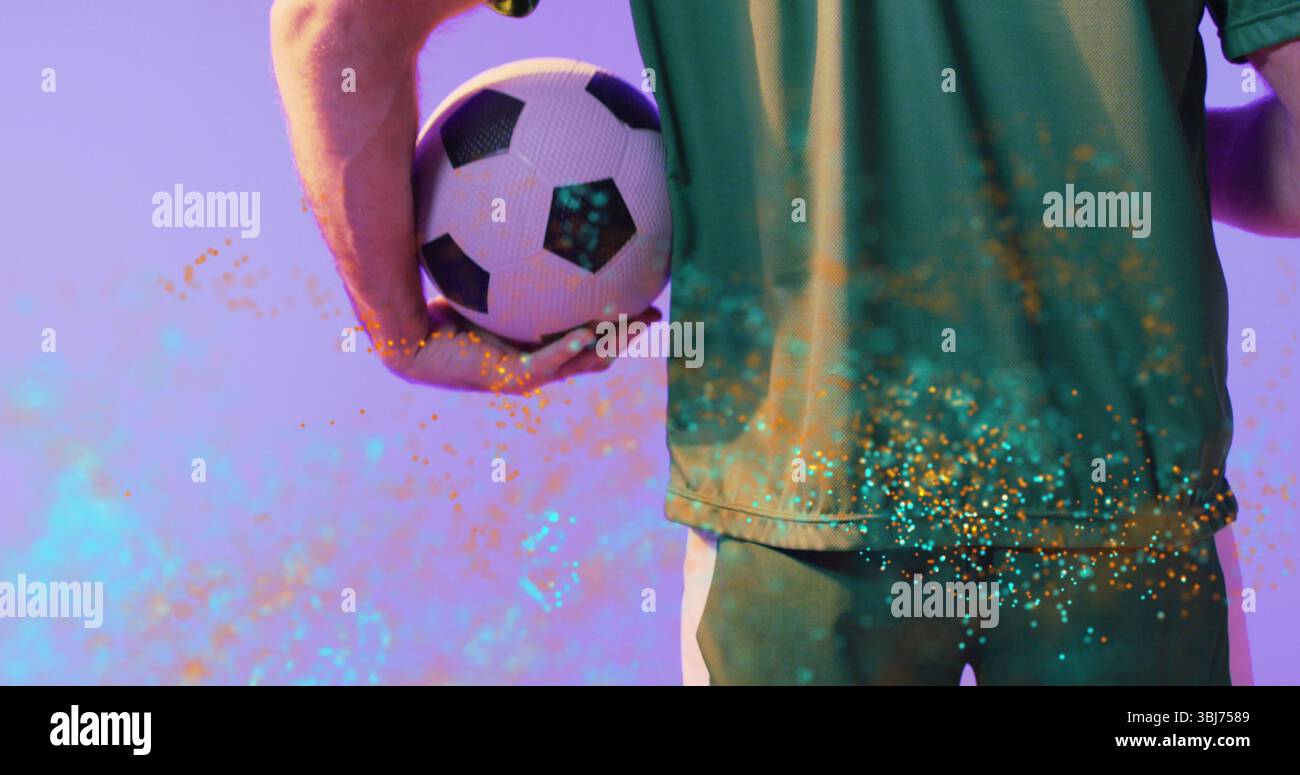 Athlet in grünem Trikot und Shorts, der im Studio Fußball hinter der Taille hält, mit Konfetti Stockfoto