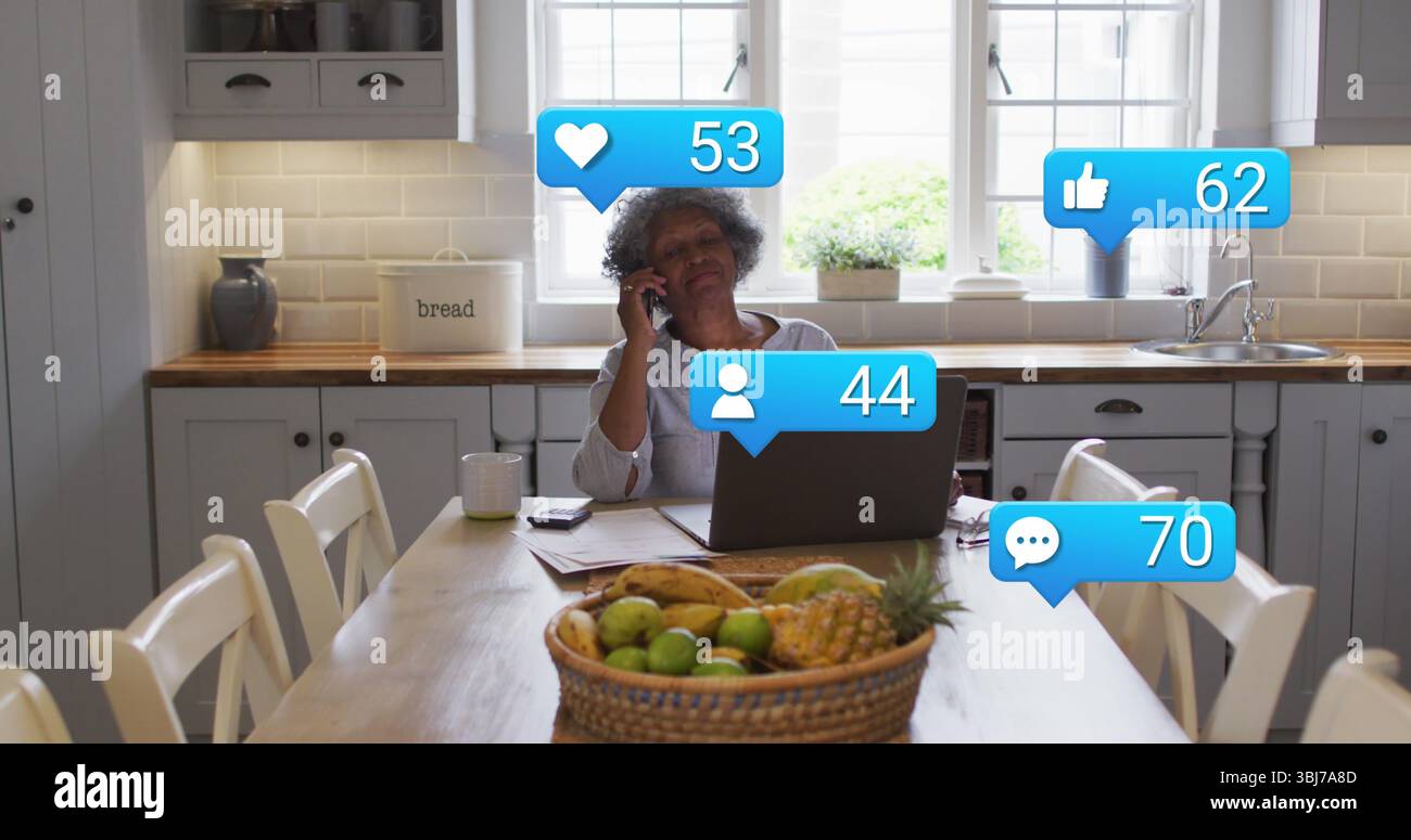 Sprechende Seniorin mit Smartphone in der modernen Küche, mit Laptop und Obstkorb Stockfoto