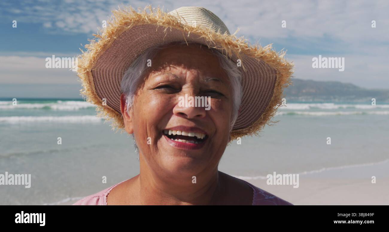 Lächelnde Seniorin mit Strohsonnenhut und rosa Bluse am Strand, mit Wellen Stockfoto