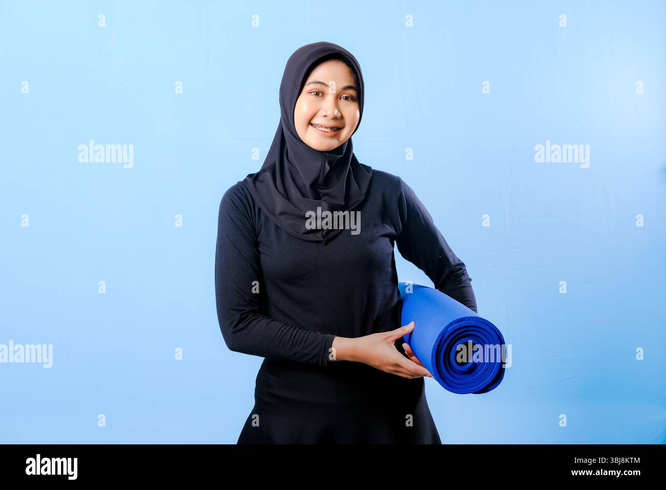 Eine Frau, die einen schwarzen Hijab trägt, hält eine blaue Yogamatte. Sie lächelt. Die Matte ist aufgerollt Stockfoto