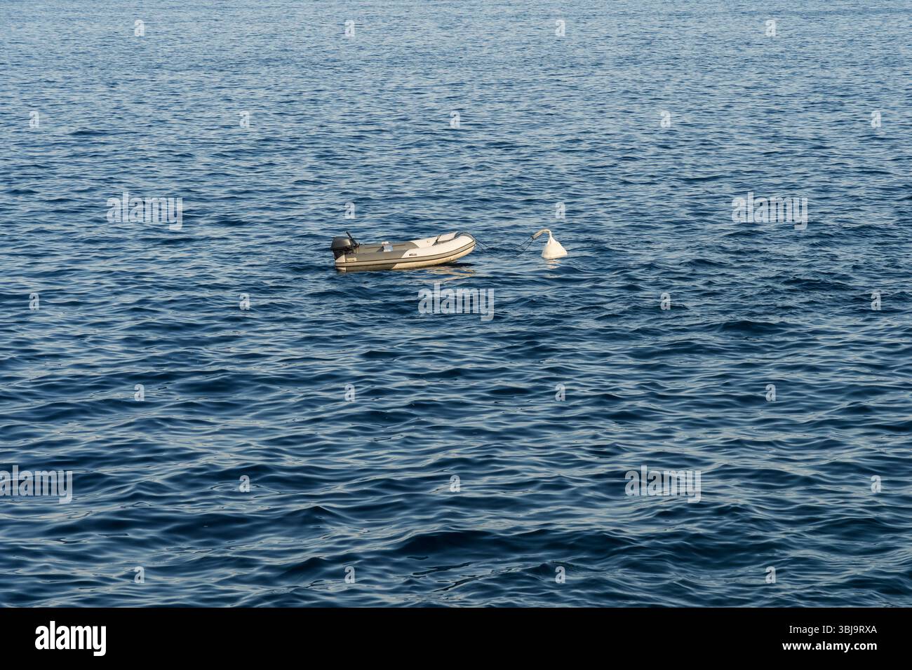 Ein kleines aufblasbares Motorboot, das an einer weißen Boje verankert ist, schwingt einsam auf den ruhigen Wellen im weiten blauen offenen Meer. Stockfoto