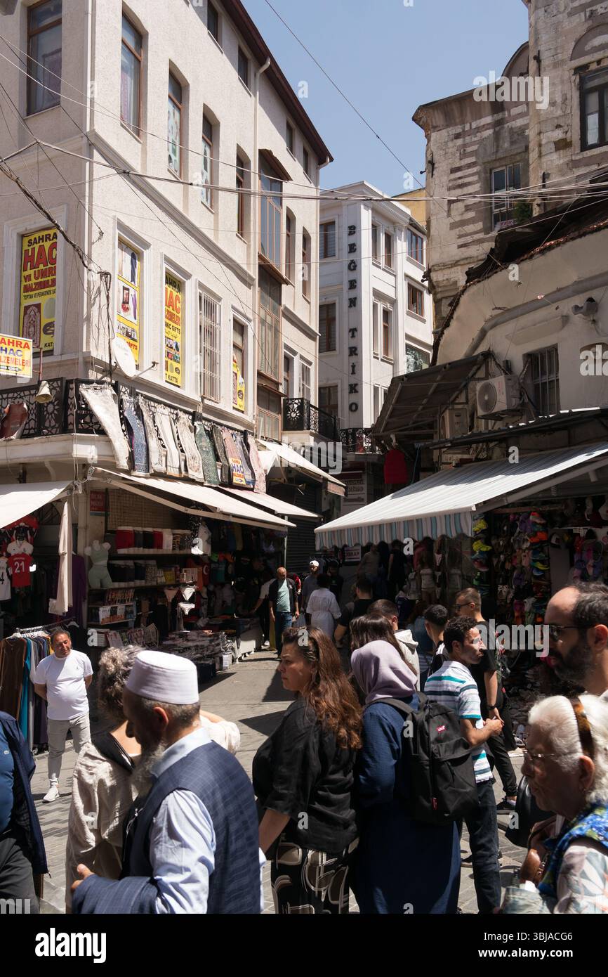 Die Menschenmassen bewegen sich auf einem lebhaften Straßenmarkt in Istanbul, umgeben von Geschäften, in denen bunte Textilien und Waren unter einem klaren blauen Himmel ausgestellt werden. Stockfoto