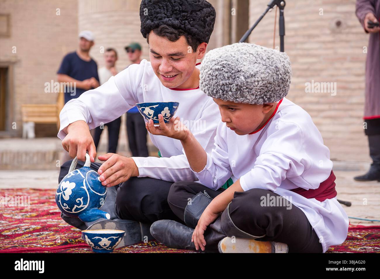 Chiwa, Usbekistan – 11. Juli 2024: Usbekische Kinder in traditioneller Kleidung während einer kulturellen Veranstaltung. Einer gießt Tee, während der andere zusieht – Darstellung von Stockfoto