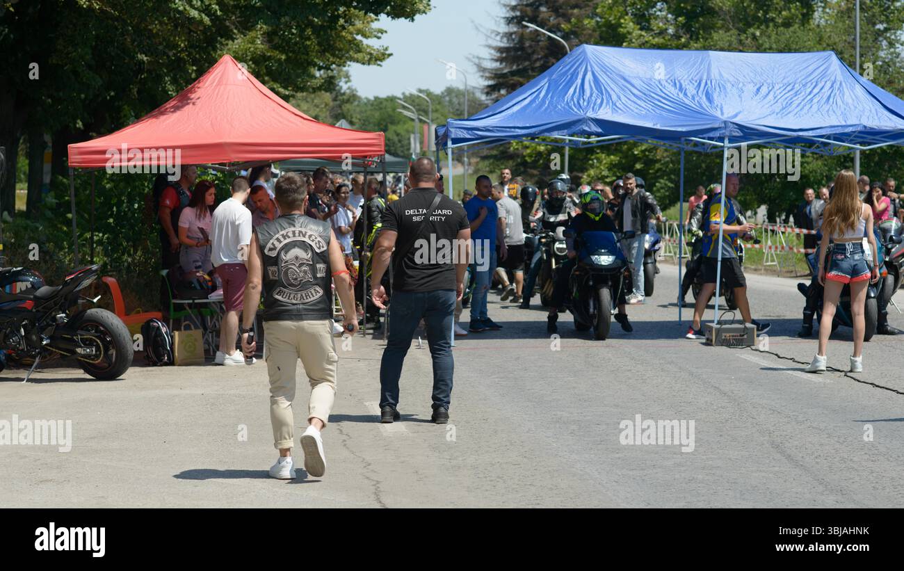 Motorradfahrer und Zuschauer treffen sich an der startlinie des Drag Race beim Moto Rock fest 2025 in der Nähe von Ruse, Bulgarien. Ein Mitglied des Sicherheitsteams von Delta Guard und ein Biker, der eine Vereinsweste von Wikings Bulgaria trägt, werden beobachtet, wie er in Richtung des aktiven Aufenthaltsbereichs läuft. Sporträder stehen unter einem blauen Zelt, während die Vorbereitungen fortgesetzt werden. Ein rotes Stützzelt und eine weibliche Starterin in Denim-Shorts sind ebenfalls sichtbar. Die Atmosphäre ist energiegeladen, während Teilnehmer und Mitarbeiter sich bei sonnigem Sommerwetter abstimmen. Stockfoto