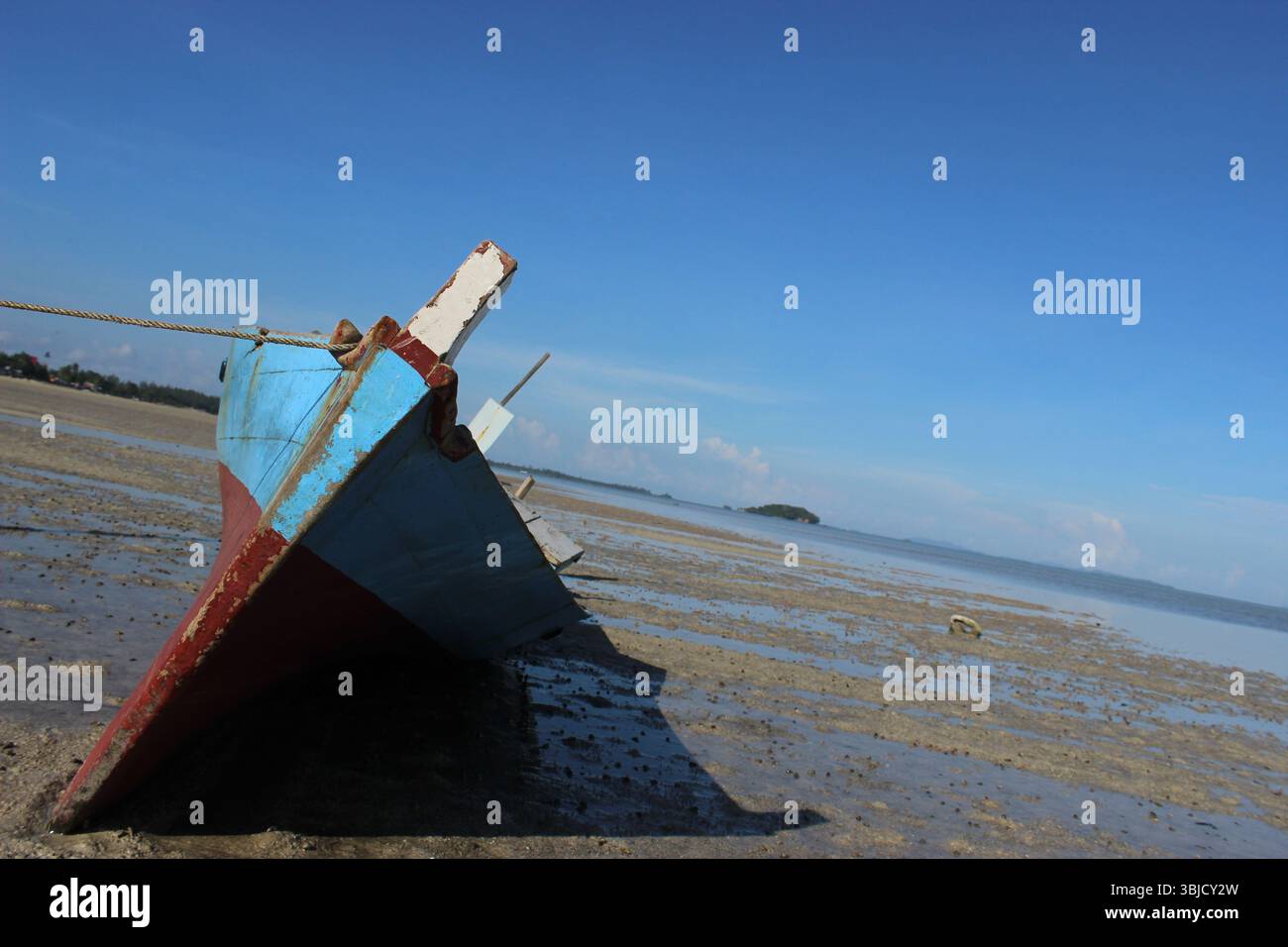 Ein traditionelles Holzboot ruht bei Ebbe auf dem exponierten Wattenmeer, mit einer ruhigen Küste im Hintergrund unter einem klaren blauen Himmel. Stockfoto