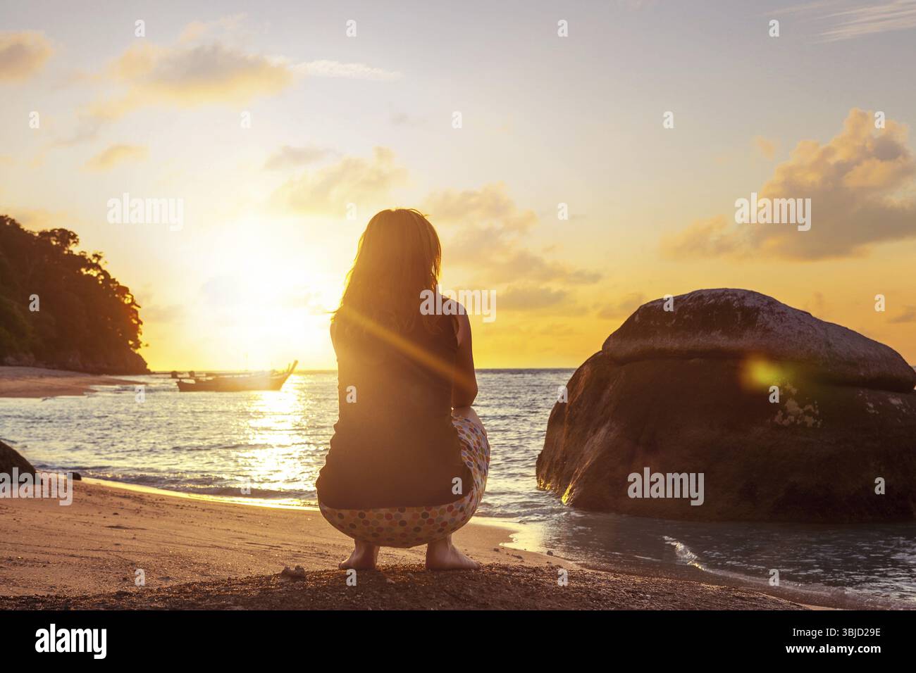 Frau, die in tiefer Hocke am Sandstrand vor dem wunderschönen Sonnenuntergang sitzt. Entspannend Stockfoto