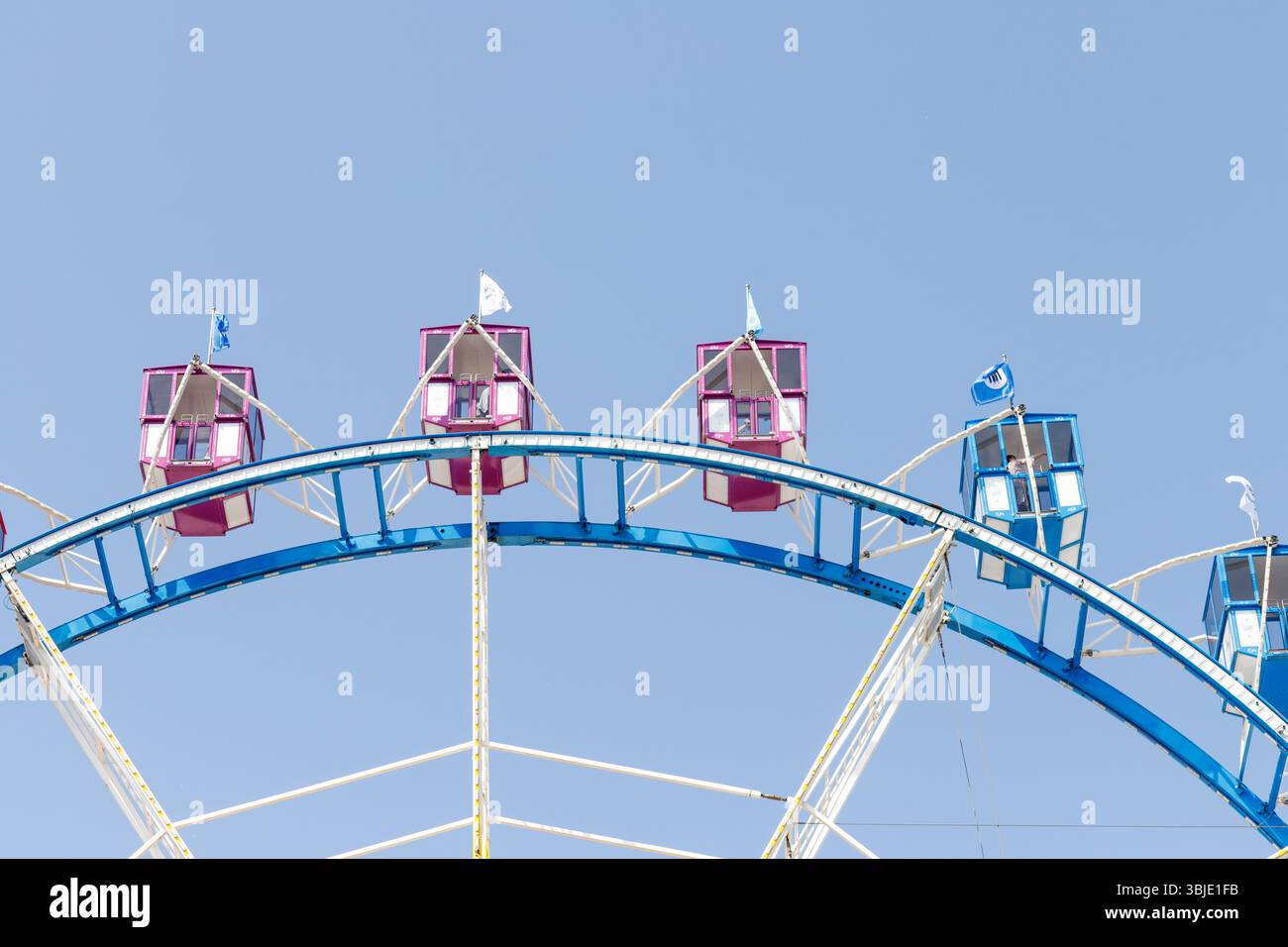 Fragment eines Riesenrads vor hellblauem Himmel mit rosa und blauen Autos und weißen Stützstrukturen. Stockfoto