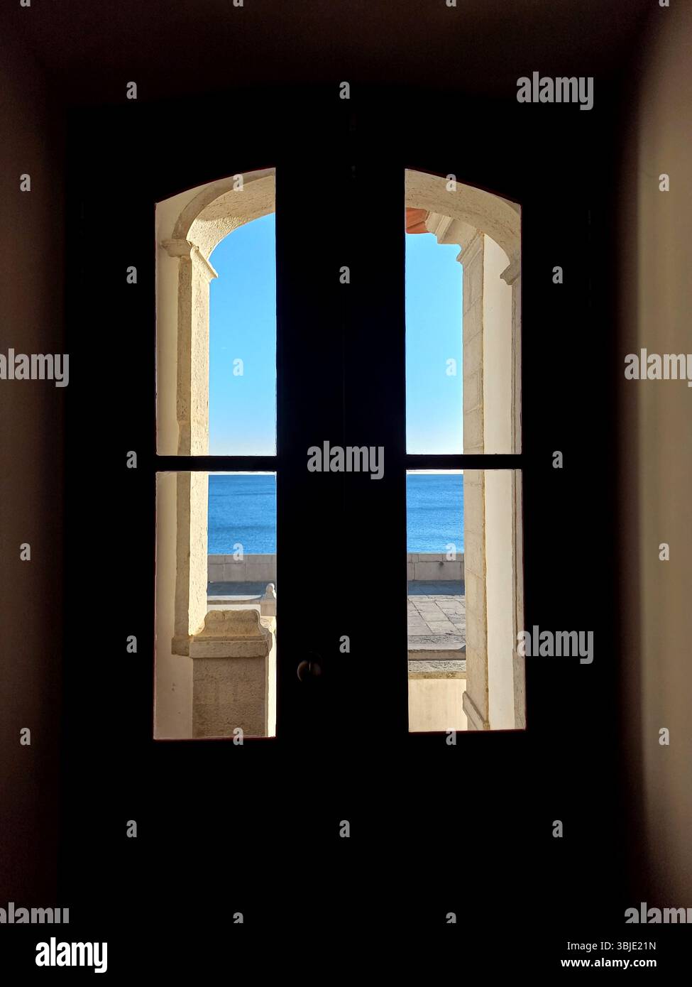 Sesimbra, Portugal. 20. Dezember 2024 Ein dunkel gerahmtes Fenster bietet einen gerahmten Blick auf den weiß getünchten Innenhof von Sesimbra, die Gebäude an den Küsten und Go Stockfoto
