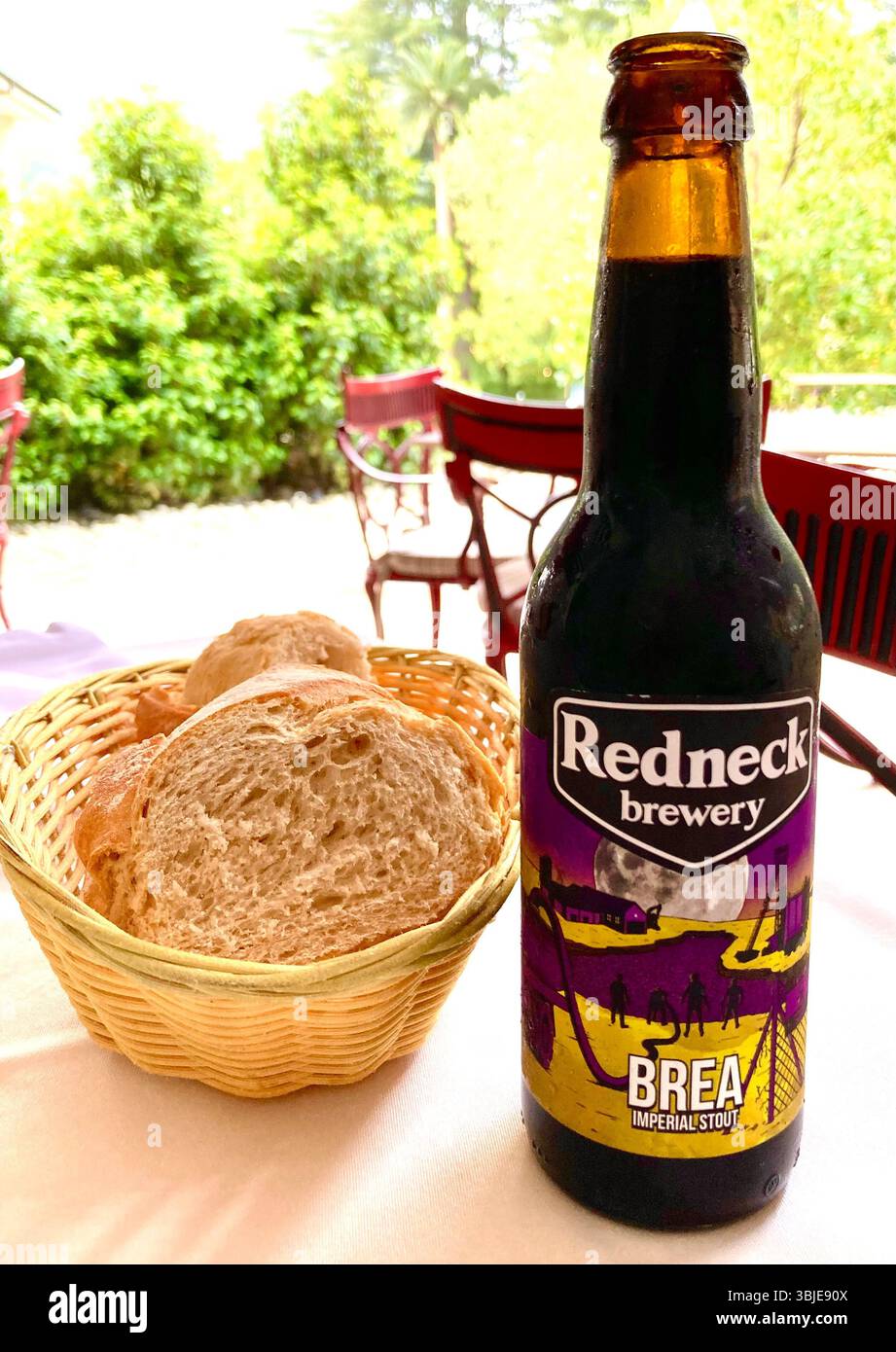 Volle Flasche kaltes Bier Brea Imperial Stout aus der spanischen Redneck-Brauerei auf einem Tisch auf der Terrasse des Restaurants mit einem Korb mit frischem Brot Stockfoto