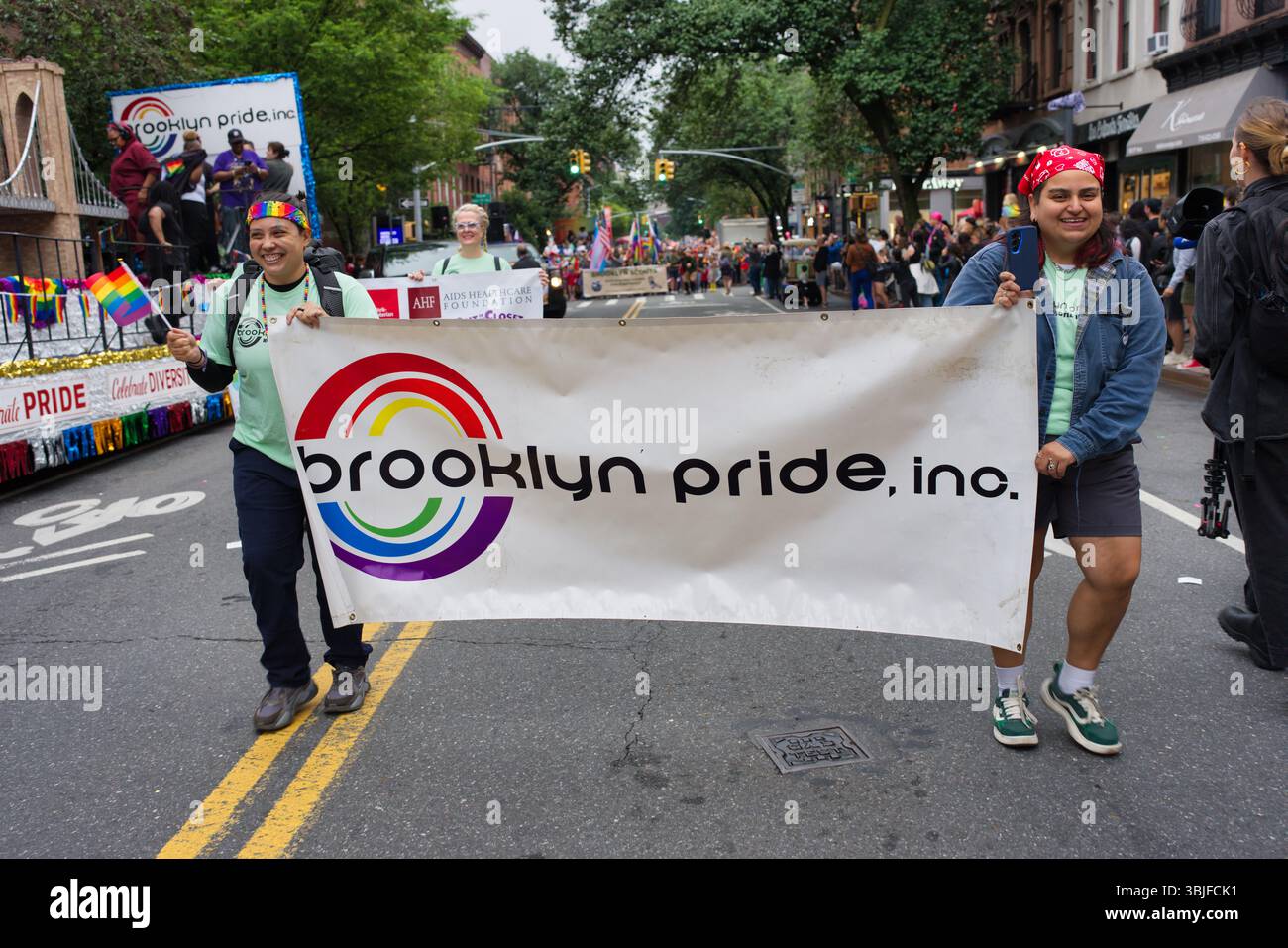 Zwei Personen halten bei einer Parade ein großes Brooklyn Pride, Inc.-Banner. Das Banner hat ein kreisförmiges Regenbogendesign. Andere Demonstranten und Zuschauer A Stockfoto