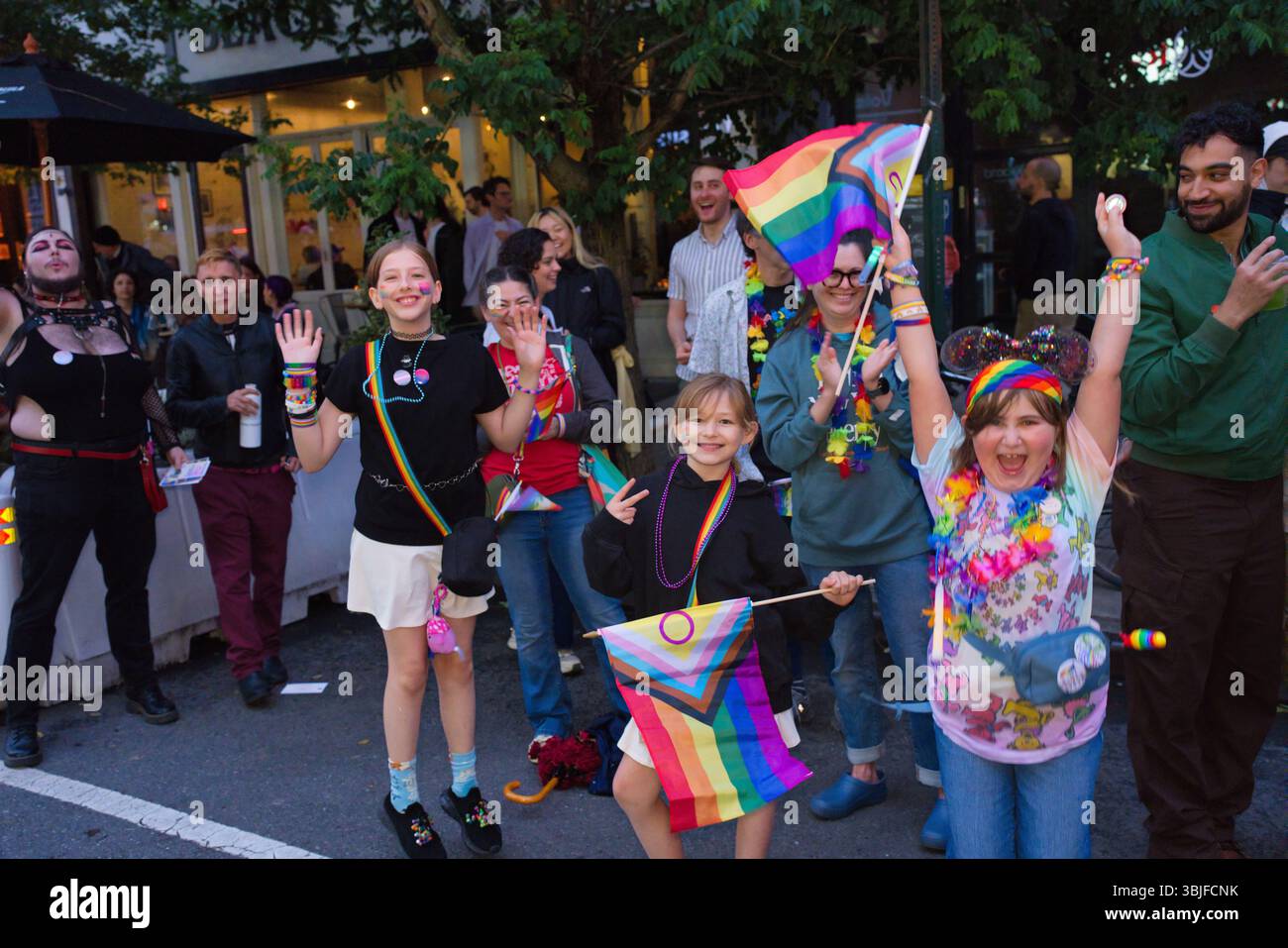 Eine vielfältige Gruppe von Menschen, viele Kinder, bei einer Pride-Veranstaltung. Sie halten Regenbogenfahnen und tragen farbenfrohe Kleidung und Accessoires. Die Szene spielt auf A Stockfoto