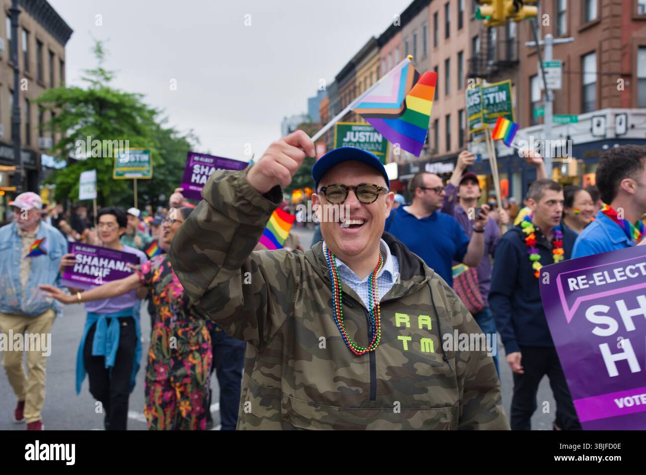 Ein lächelnder Mann in Tarnjacke hält bei einer politischen Parade eine Regenbogenfahne. Andere Demonstranten halten Schilder, die verschiedene Kandidaten unterstützen. Der Cit Stockfoto