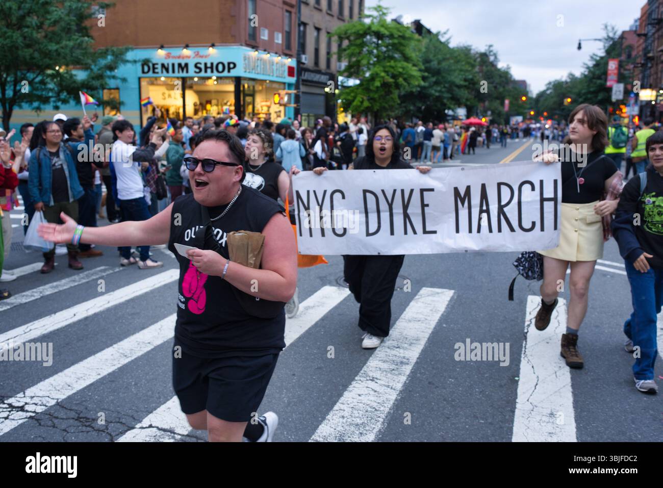 Eine fröhliche Straßenszene fängt den NYC Dyke March ein. Eine Person in einem schwarzen Hemd und einer kurzen Hose führt die Prozession an, während andere ein Banner tragen, das ausruft Stockfoto