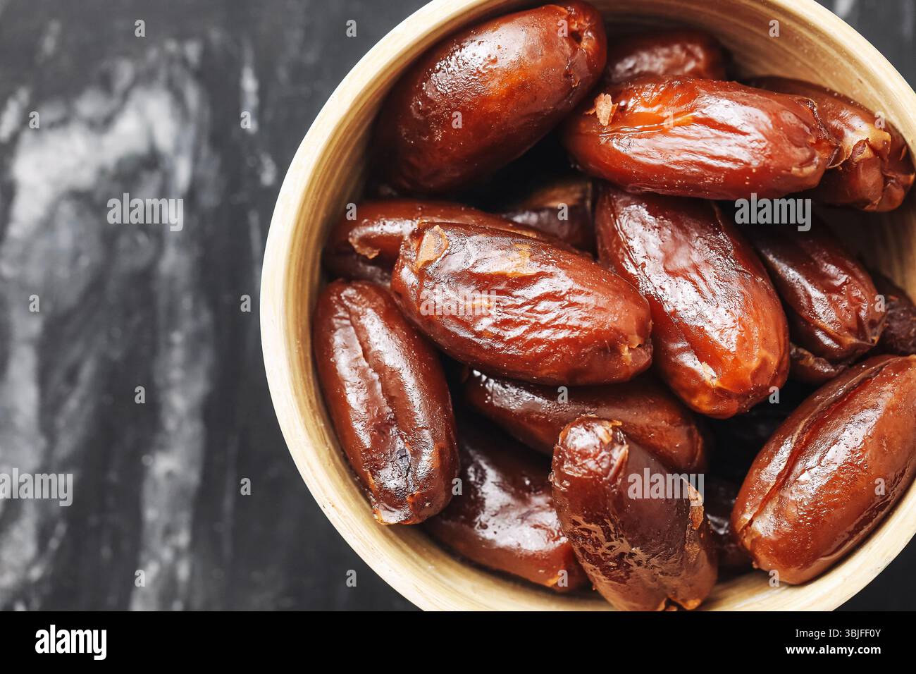 Datteln in Holzschale. Süße getrocknete Früchte. Bio-gesunder Snack. Natürliche Zuckerquelle. Braune glänzende Textur. Nahaufnahme auf schwarzem Marmor. Stockfoto