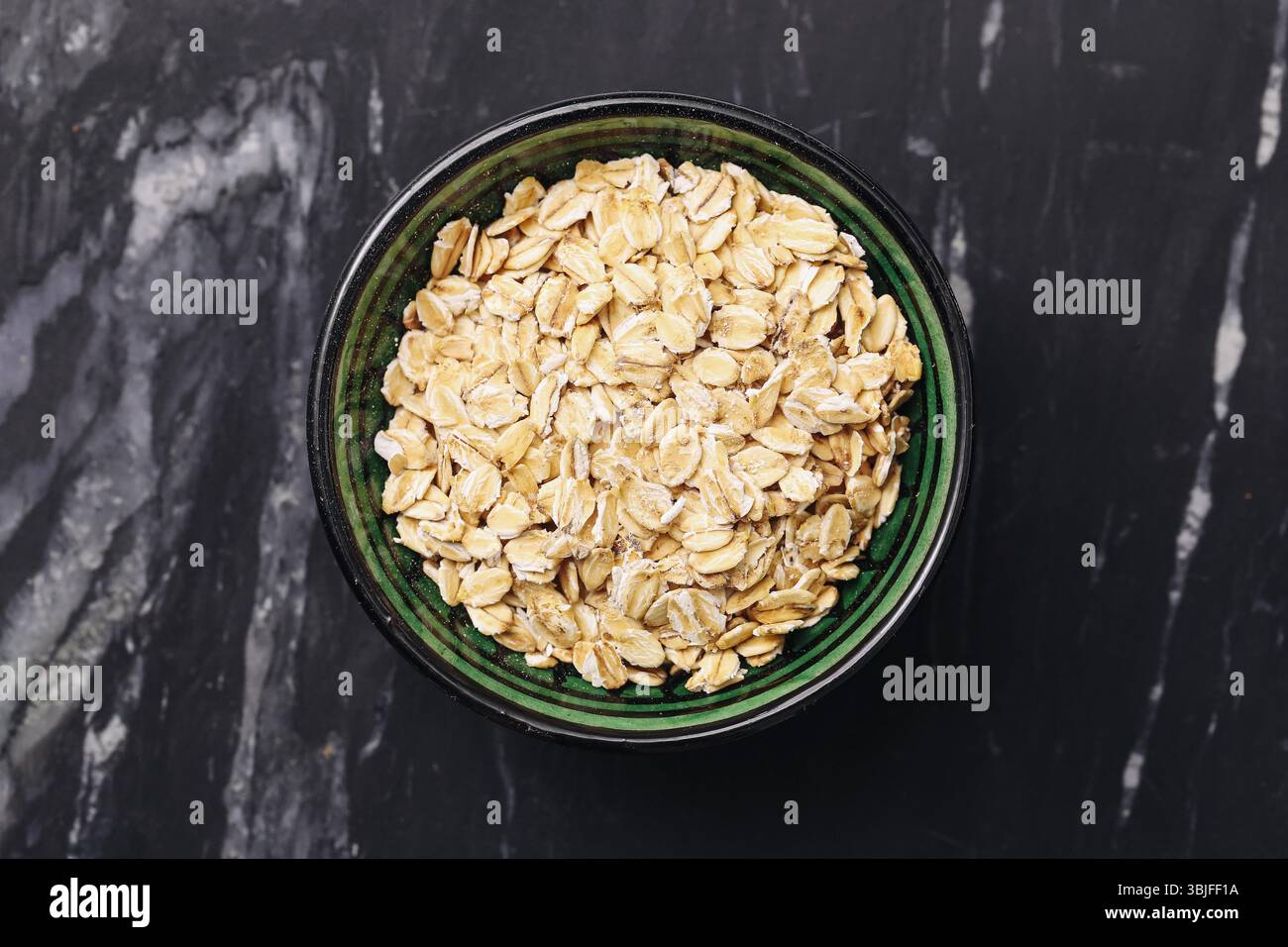 Haferflocken in der Schüssel. Gesunde Frühstücksidee. Trockenes Müsli auf dunklem Marmor. Bio-Vollkornhafer. Nahrhafte Morgenmahlzeit. Einfache, saubere Zutaten. Stockfoto