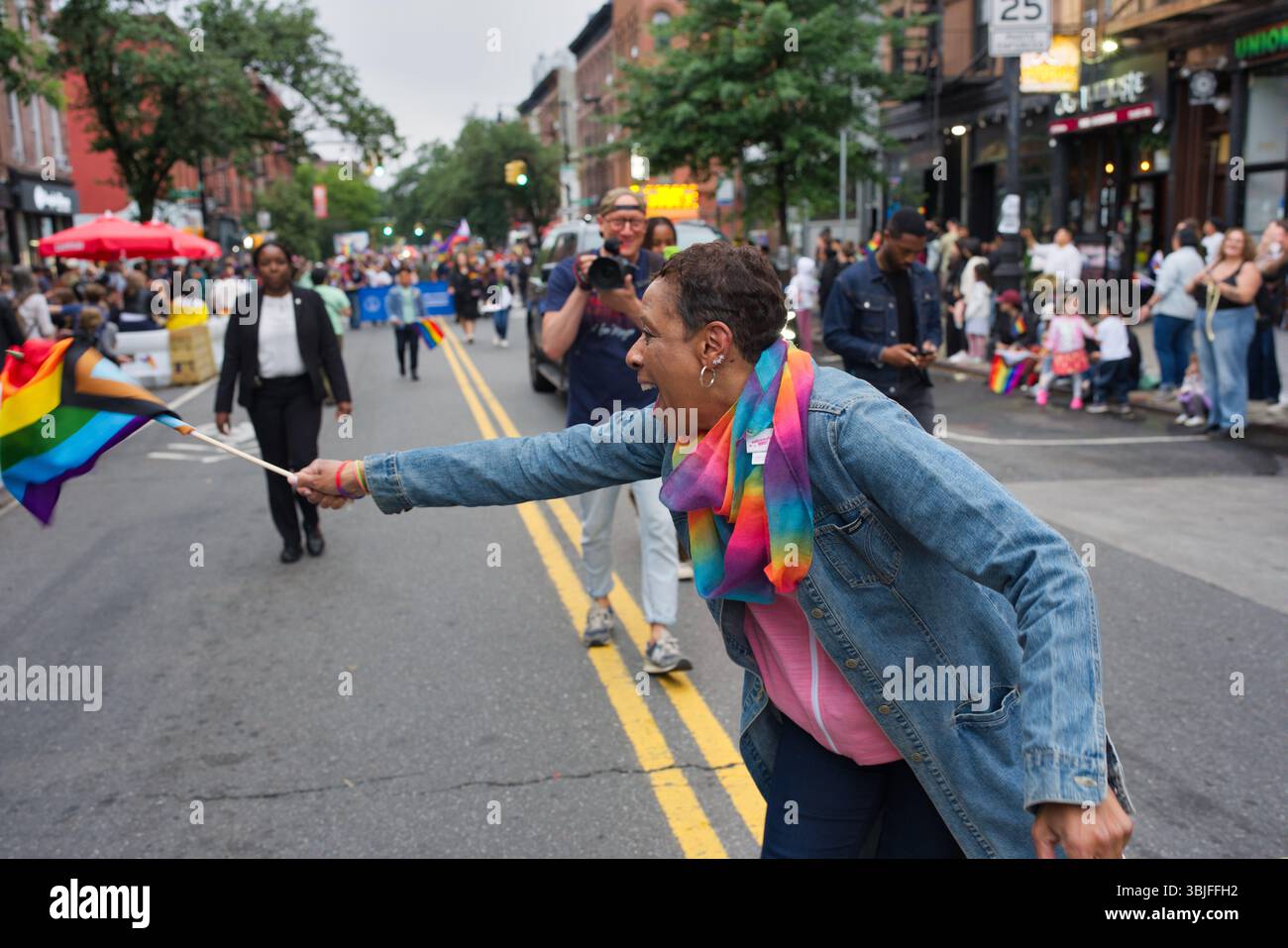 Eine Frau schwingt begeistert eine Regenbogenfahne bei einer Pride-Parade. Sie trägt eine Jeansjacke und einen Regenbogenschal, umgeben von anderen Paradeparaden Stockfoto