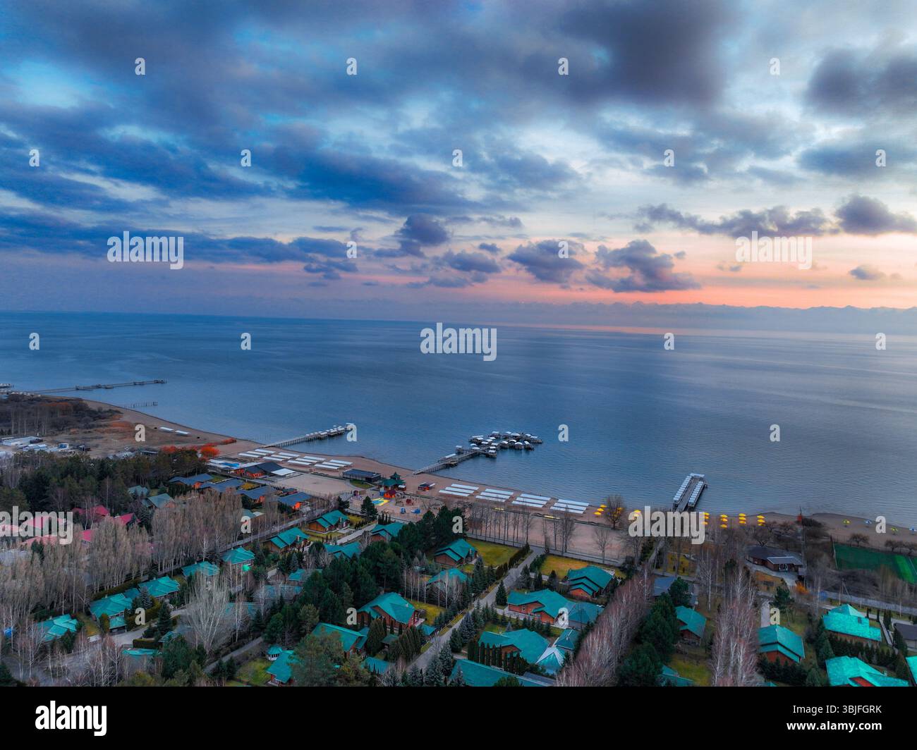 Aus der Vogelperspektive auf den Issyk Kul See unter einem bedeckten Sonnenuntergang, der einen Küstenort mit Gebäuden zwischen Bäumen und ruhigem Seenwasser zeigt Stockfoto