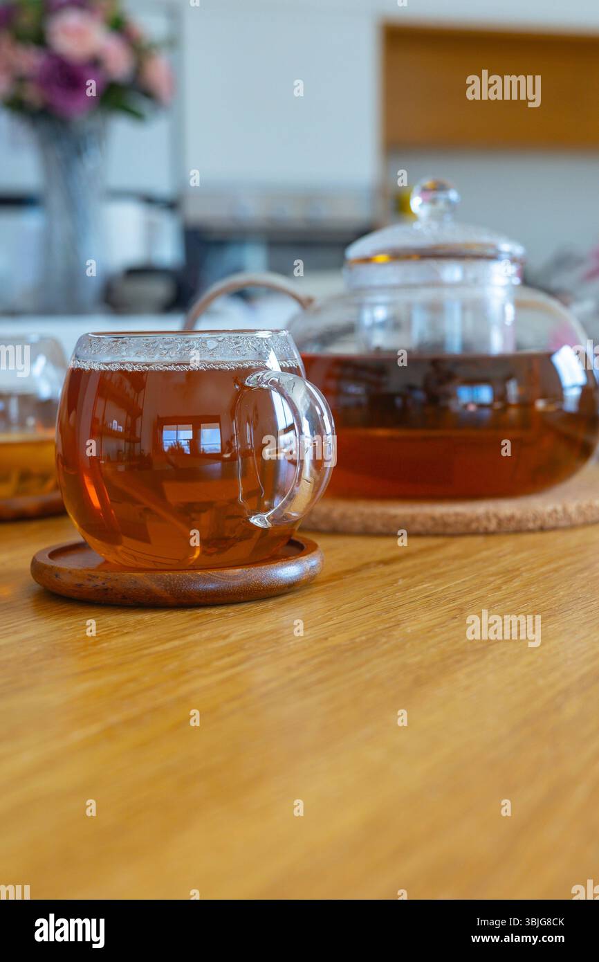 Teekanne aus Glas und Tassen mit heißem Tee und Zitrone auf Holztisch im hellen Wohnzimmer. Warmes Licht und gemütliches Interieur. Konzept von Wellness und Entspannung Stockfoto