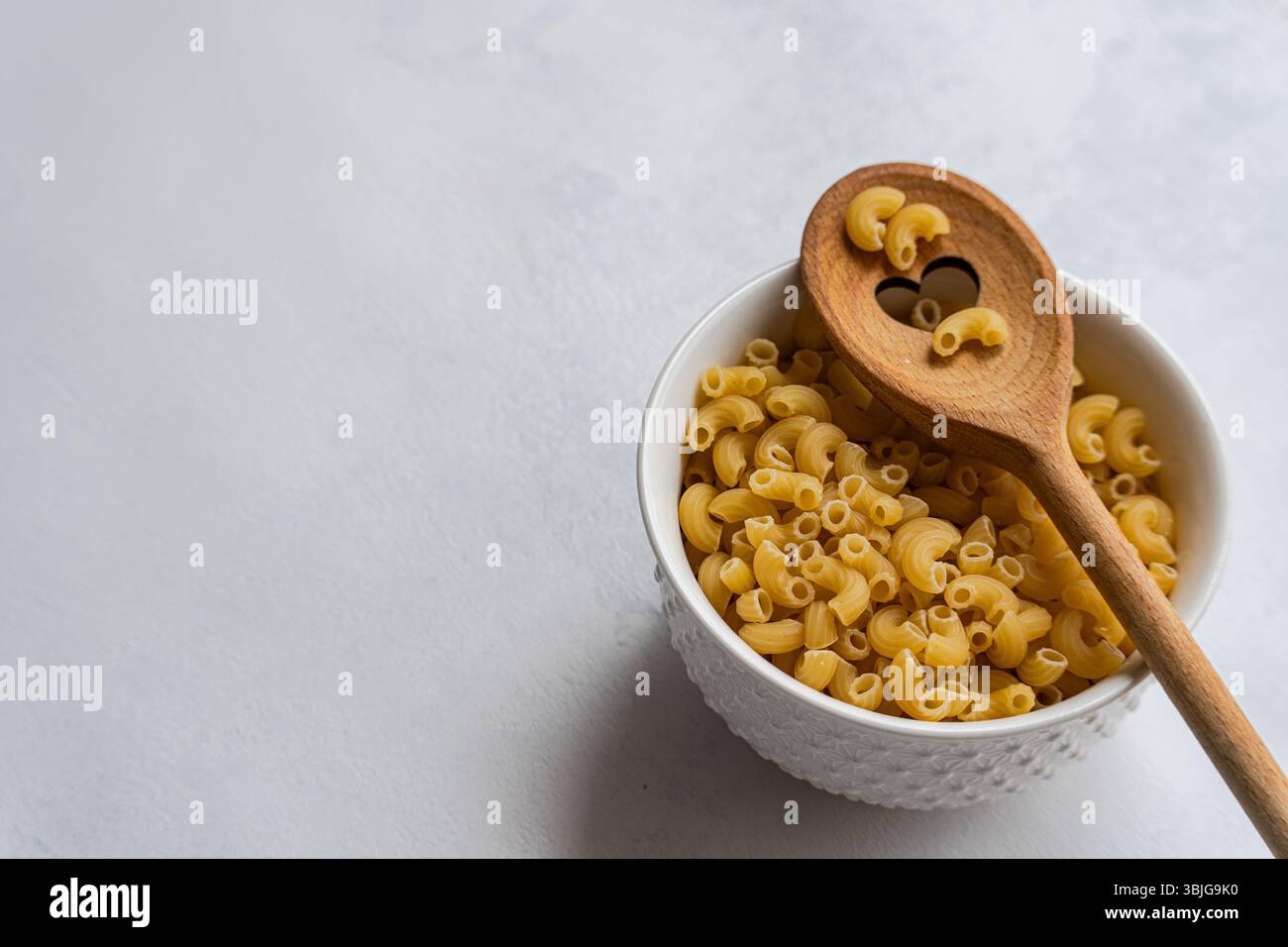 Nahaufnahme einer Schüssel mit roher Makkaroni-Pasta und einem hölzernen Löffel mit Herzform auf einer Küchenarbeitsfläche Stockfoto