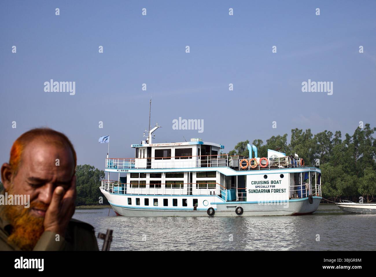 Ein Waldhüter mit Henna-gefärbtem Bart ruht, während das ML Bon Bibi Boot, das für Sundarbans Touren und Patrouillen genutzt wird, an den Mangrovenbänken vorbeischwimmt. Stockfoto