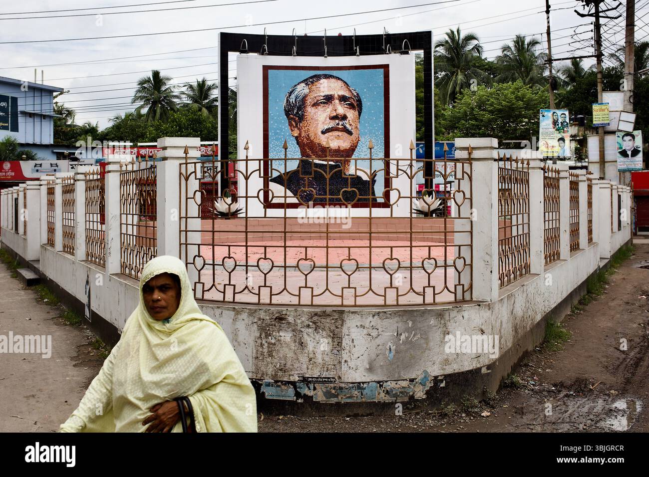 Das Bangabandhu Smrity Mural in der Stadt Jessore durch kunstvolle Zäune, mit einer Frau in einem cremefarbenen Tuch, die im Vordergrund vorbeigeht. Stockfoto