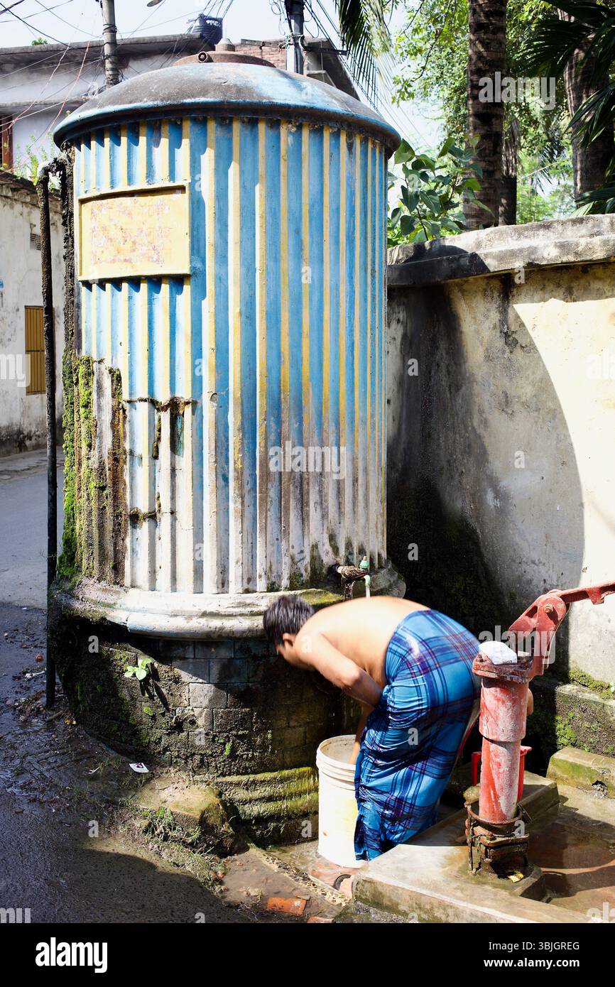 Der Mann wäscht sich an einer Straßenpumpe neben einem Dhopkol-Wassertank in Rajshahi, Bangladesch, und veranschaulicht die tägliche Abhängigkeit vom öffentlichen Wasserzugang. Stockfoto