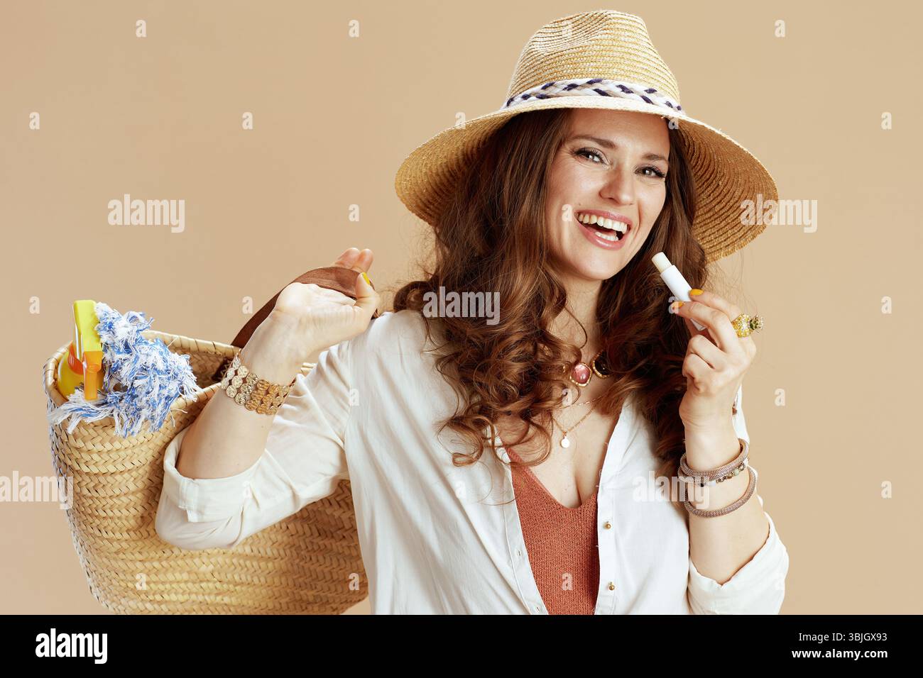 Strandurlaub. Glückliche stilvolle Frau in weißer Bluse und Shorts isoliert auf beigefarbenem Hintergrund mit Strohbeutel, hygienischem Lippenstift und Strohhut. Stockfoto Strandurlaub. Glückliche stilvolle Frau in weißer Bluse und Shorts isoliert auf beigefarbenem Hintergrund mit Strohbeutel, hygienischem Lippenstift und Strohhut. Stockfoto