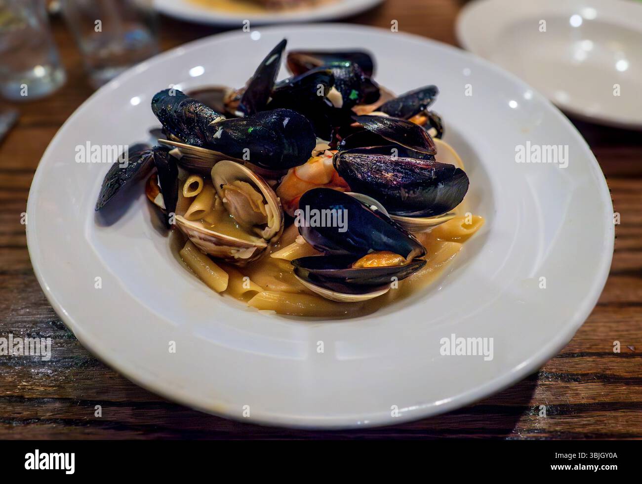 Eine weiße Platte ist voller Zuppa di Pesce – dicke Muscheln, Muscheln und Garnelen auf der Penne, getränkt mit einer herzhaften Wein- und Buttersauce, die das Essen fängt Stockfoto