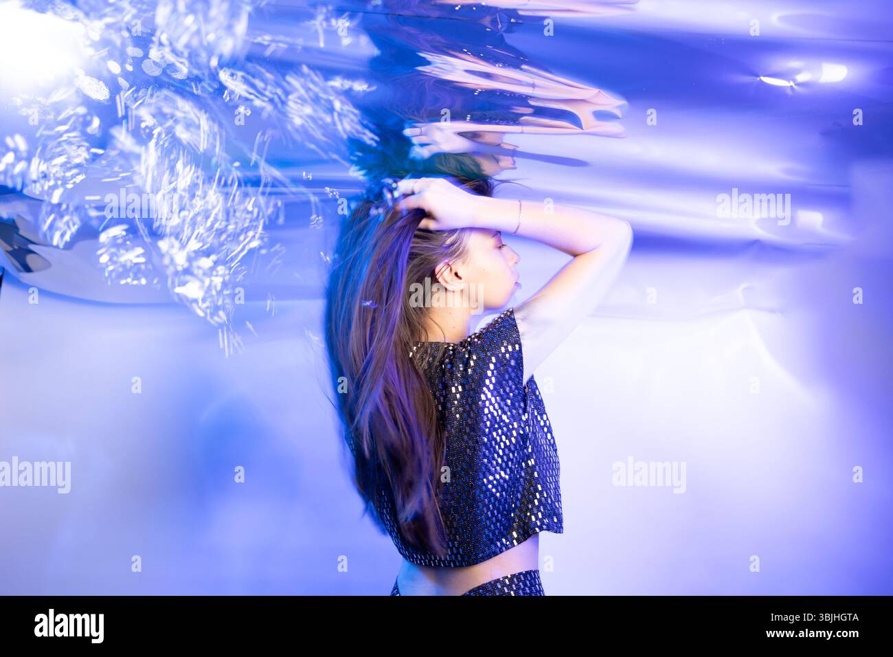 Eine Frau posiert unter Wasser mit ihren Haaren, die sich anmutig um sie herum bewegen. Das Umgebungslicht erzeugt schimmernde Reflexionen und verleiht der ruhigen Atmosphäre einen künstlerischen Touch. Stockfoto