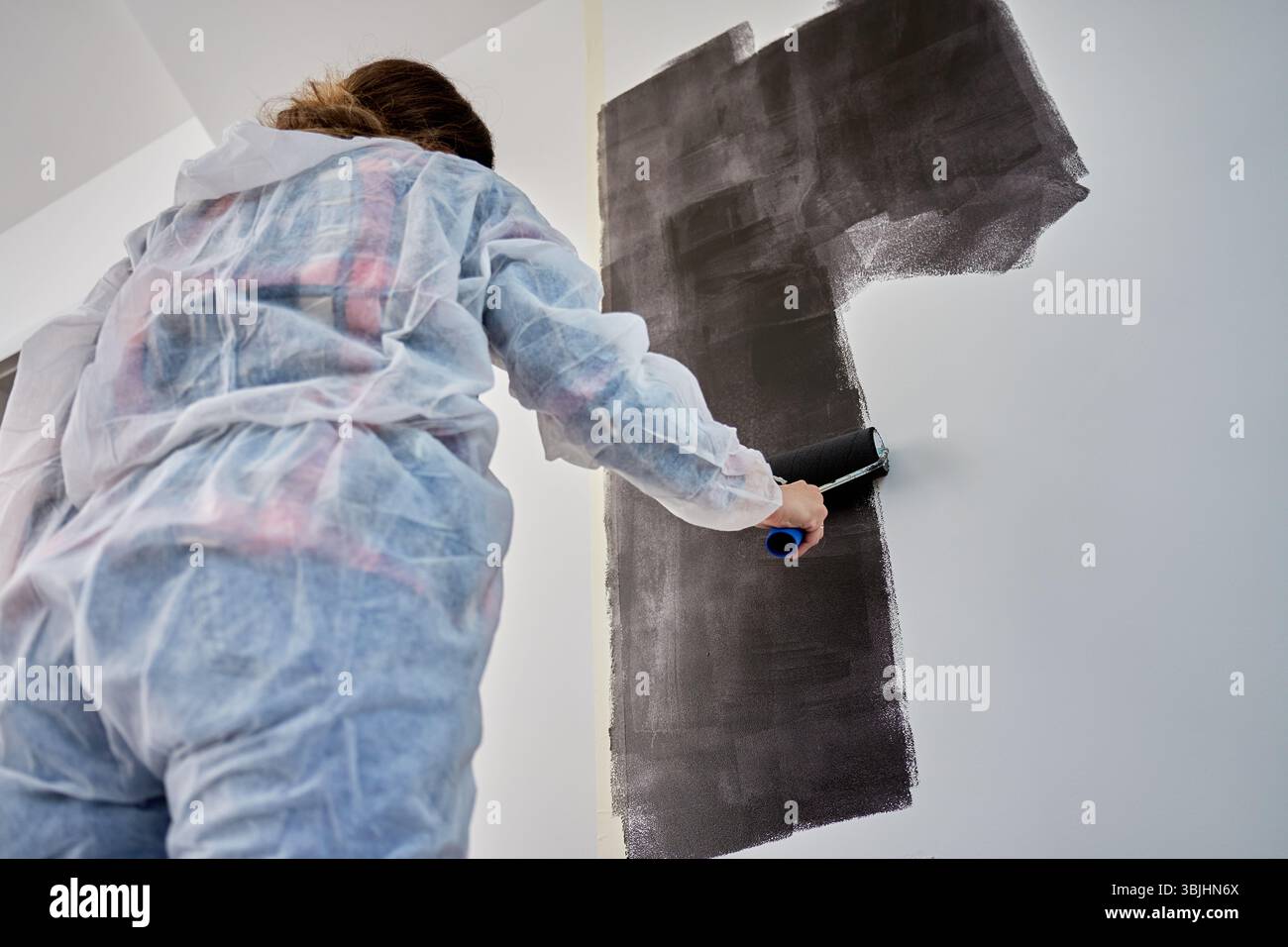 Frau in Schutzanzug Hose weiße Wand mit schwarzem Druck mit Rolle während der Renovierung. Weibliche Arbeiterin, die die Innenwand anstrich. Konzept des Home Impro Stockfoto