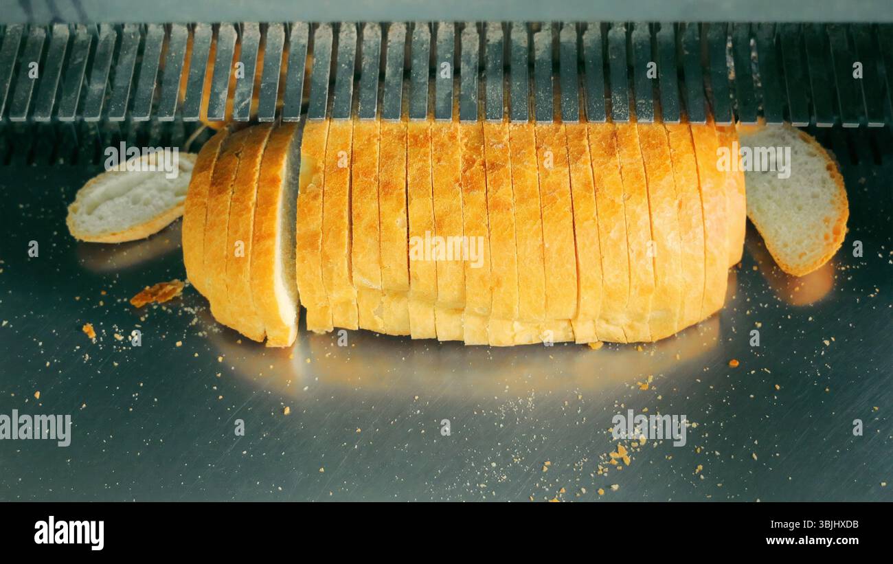 Frisch Gebackener Brotbrot Wird In Scheiben Geschnitten. Eine Nahaufnahme eines frisch gebackenen Brotlaibs, der von einer Brotschneidemaschine geschnitten wird, mit verstreuten Krümeln. Stockfoto