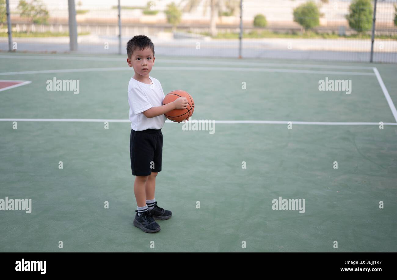 Der 4-jährige Junge steht mit einem Ball auf dem Basketballfeld und zeigt Konzentration und Bereitschaft, betont die Bedeutung des Schwerpunkts im Sport und Stockfoto