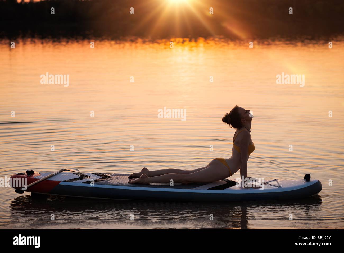 Frau, die beim Sonnenuntergang auf dem Paddleboard steht und dabei ruhiges Wasser, goldenes Licht und achtsames Training im Freien genießt. Stockfoto