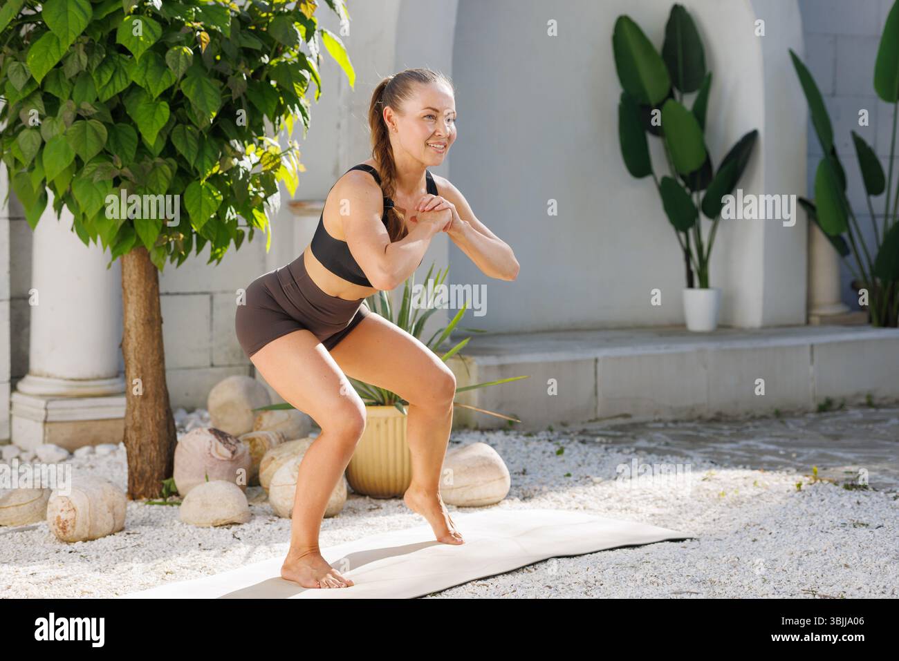 Frau in schwarzer Sportbekleidung, die Squat-Übungen im Freien macht, Stärke, Motivation und gesunden Lebensstil zeigt. Stockfoto