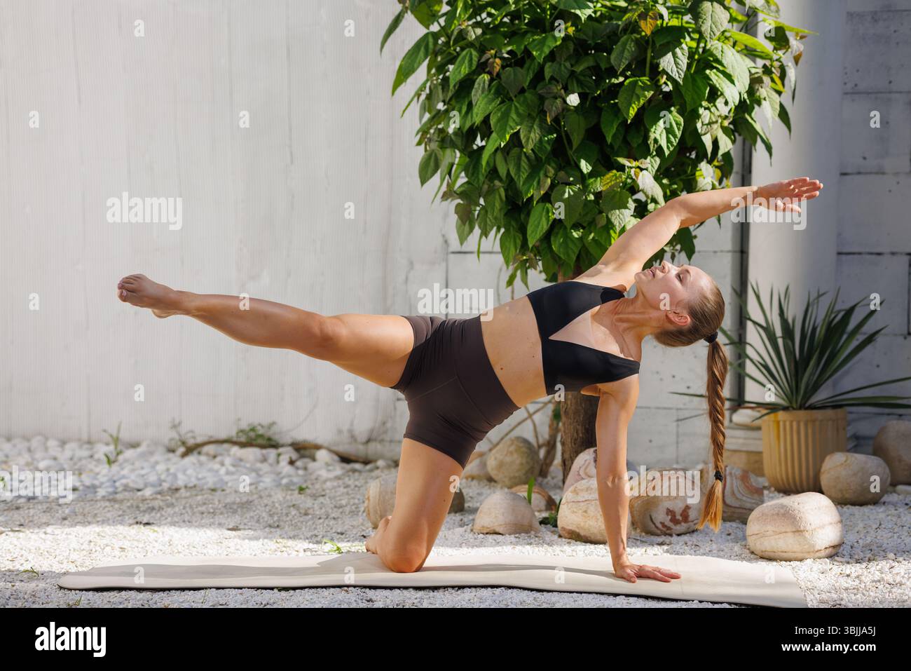 Frau in schwarzer Sportbekleidung balanciert in einer Yoga-Pose im Freien, zeigt Stärke, Flexibilität und konzentriert sich auf eine Matte. Stockfoto