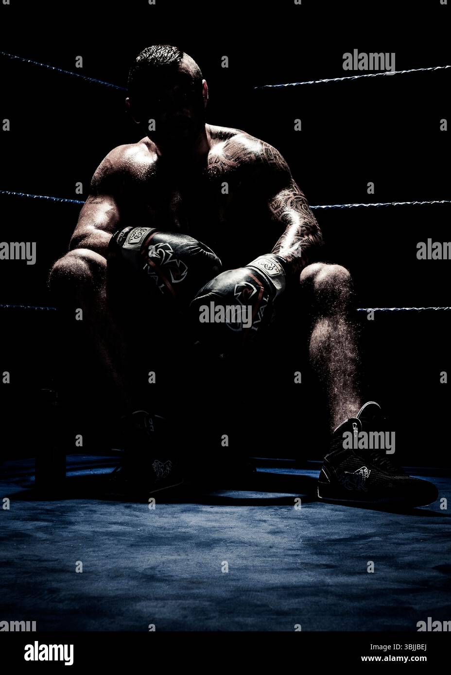 Ein Boxer sitzt im Ring, beleuchtet von einem Scheinwerfer. Er scheint sich einen Moment zu nehmen, vielleicht über ein Spiel nachzudenken. Stockfoto